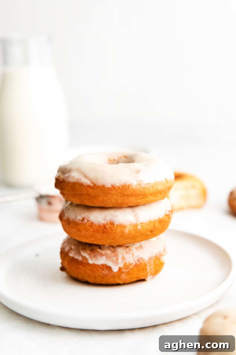 Three beautifully glazed old-fashioned buttermilk doughnuts stacked on top of one another on a pristine white plate, with a clear glass milk jug filled with milk in the soft-focus background.