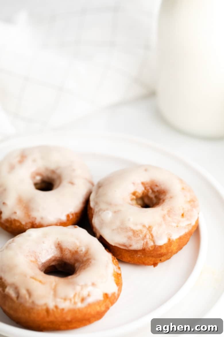 Three perfectly glazed old-fashioned doughnuts on a white plate, with a rustic glass container of milk beside them, ready to be enjoyed as a classic breakfast.