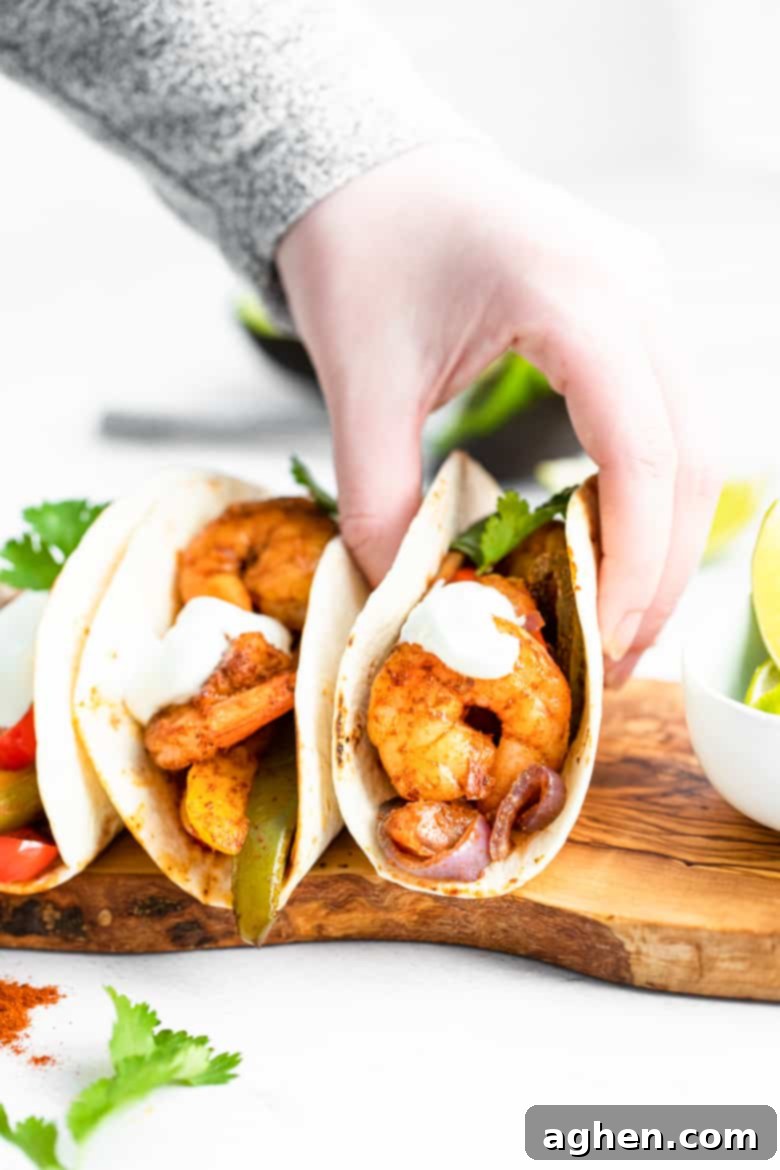 Easy Sheet Pan Shrimp Fajitas 3 Hand picking up a warm tortilla filled with seasoned shrimp, tender peppers, sweet onions, and a dollop of sour cream, ready for a bite.