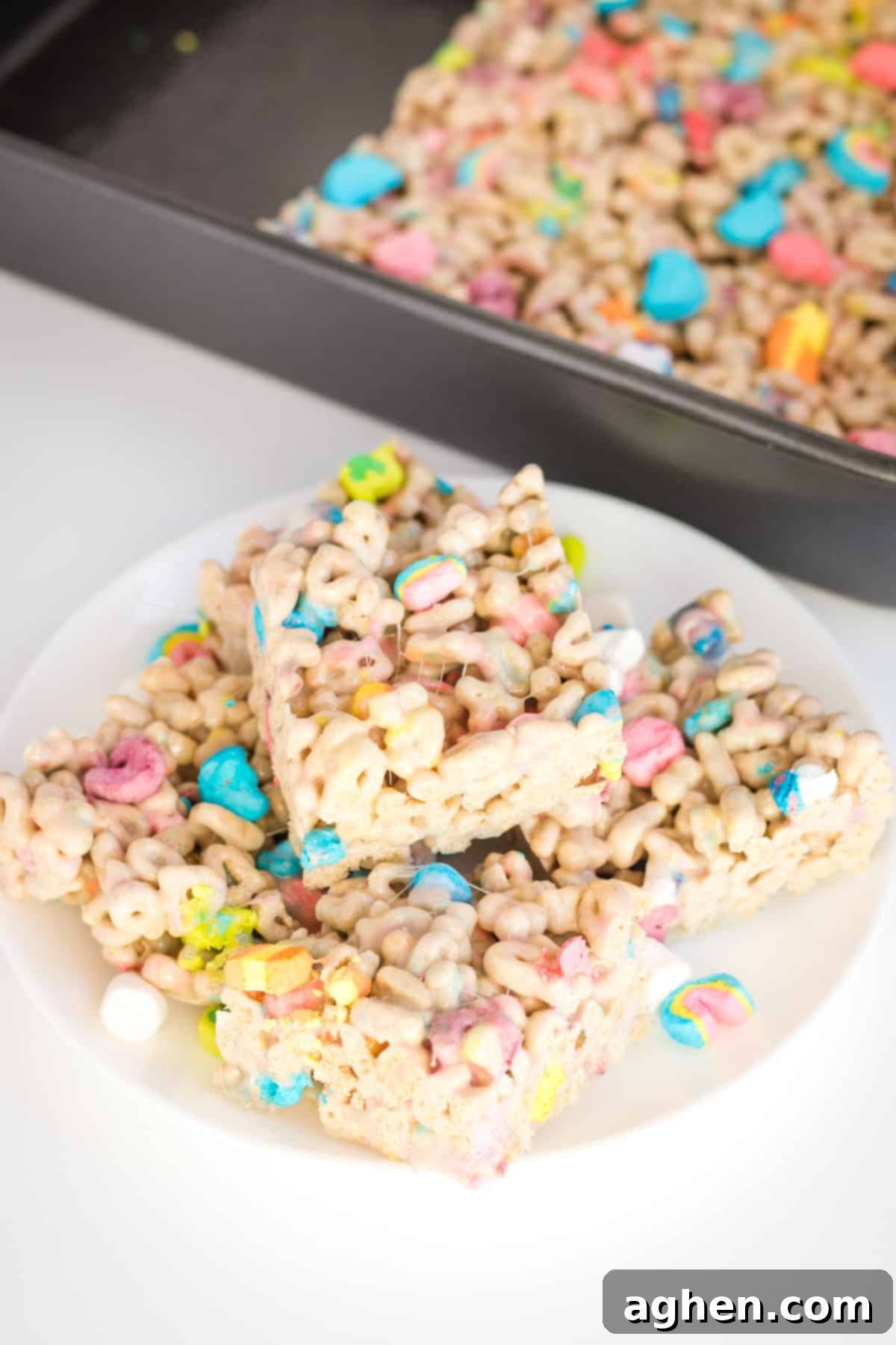 Lucky charms bars cut into squares and piled on a white plate. Baking dish with uncut treats is behind them.