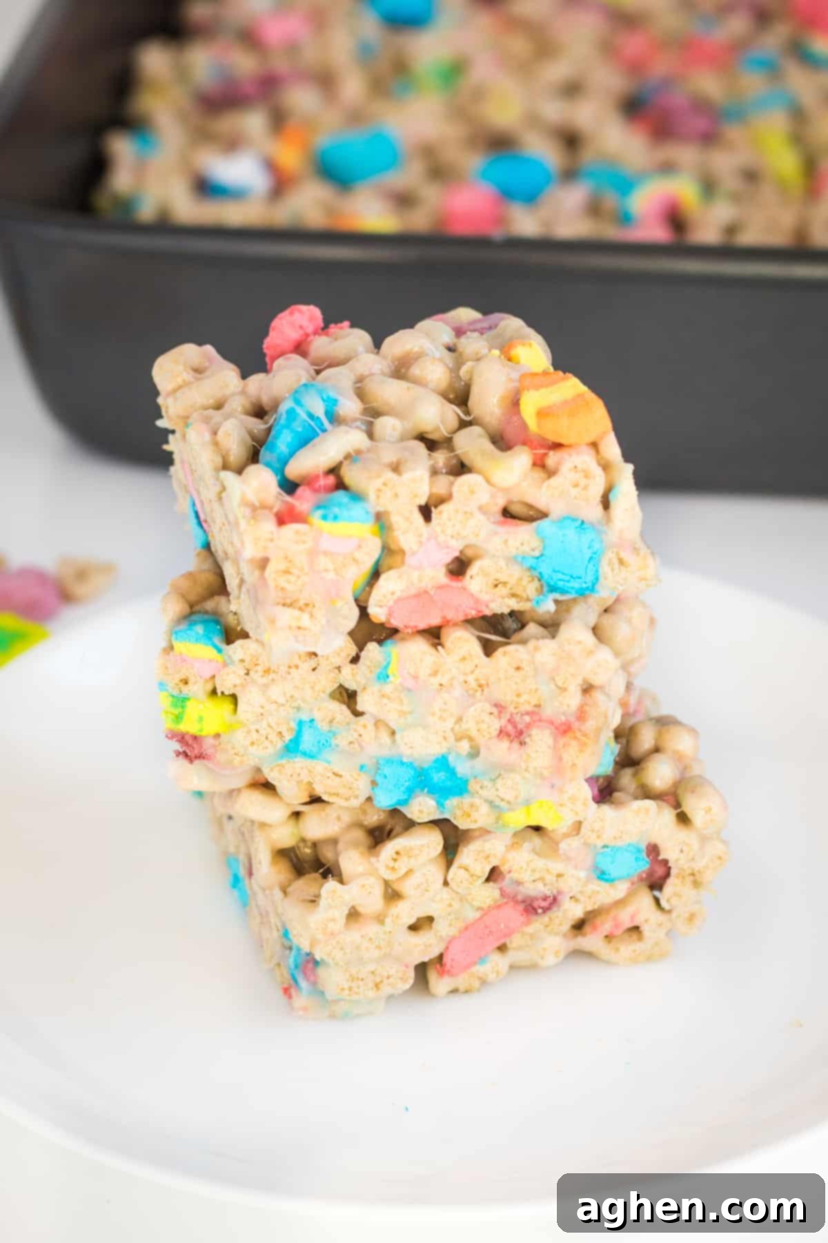 Three lucky charms marshmallow cereal bars cut into squares and stacked on top of one another on a white plate. A baking pan with uncut treats is behind them.