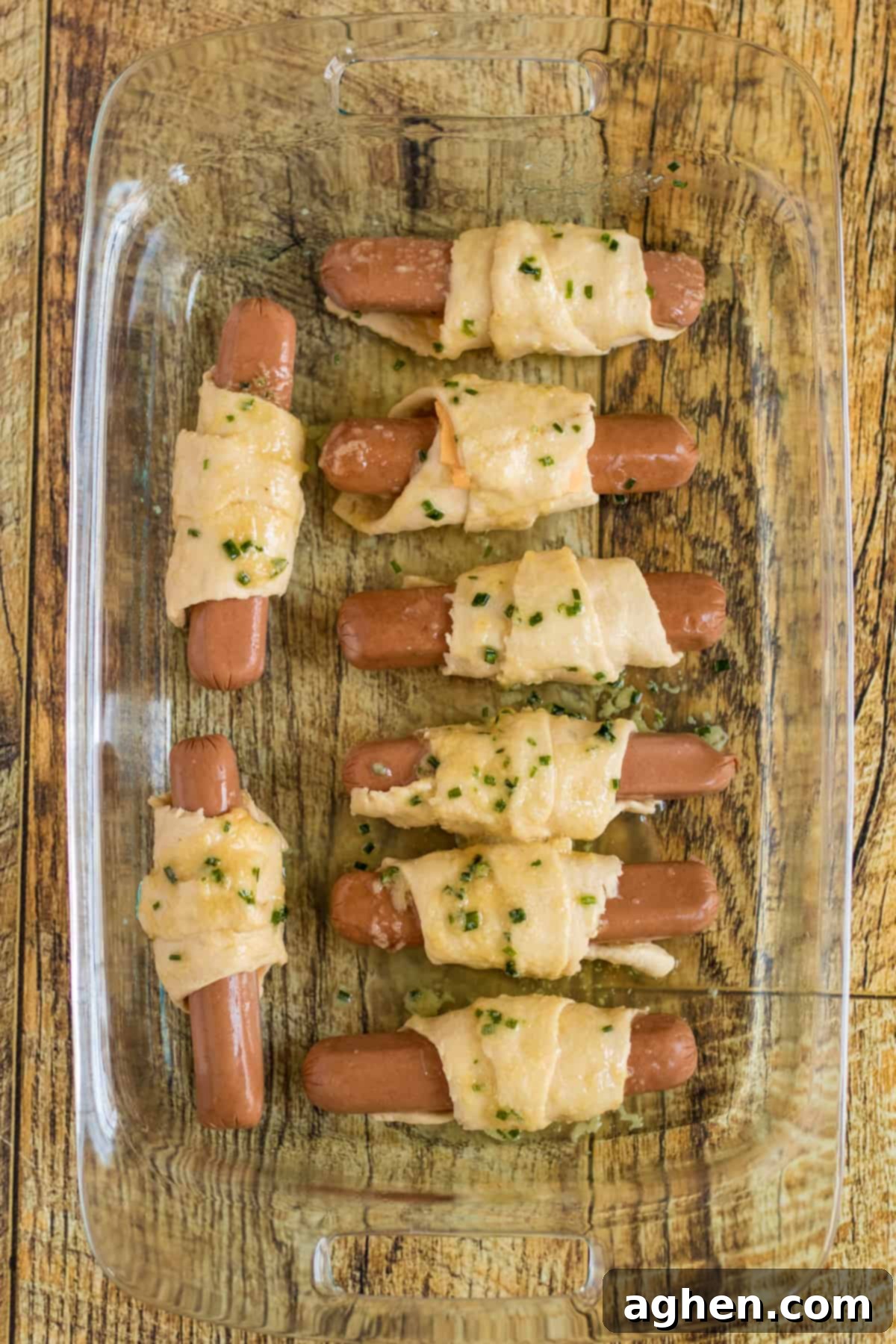 Unbaked crescent-wrapped hot dogs arranged in glass baking dish and topped with garlic and chive butter