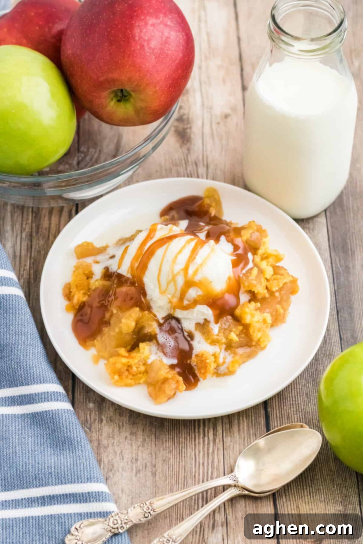 Apple dump cake topped with vanilla ice cream and caramel sauce. Glass of milk and apples are next to plate.