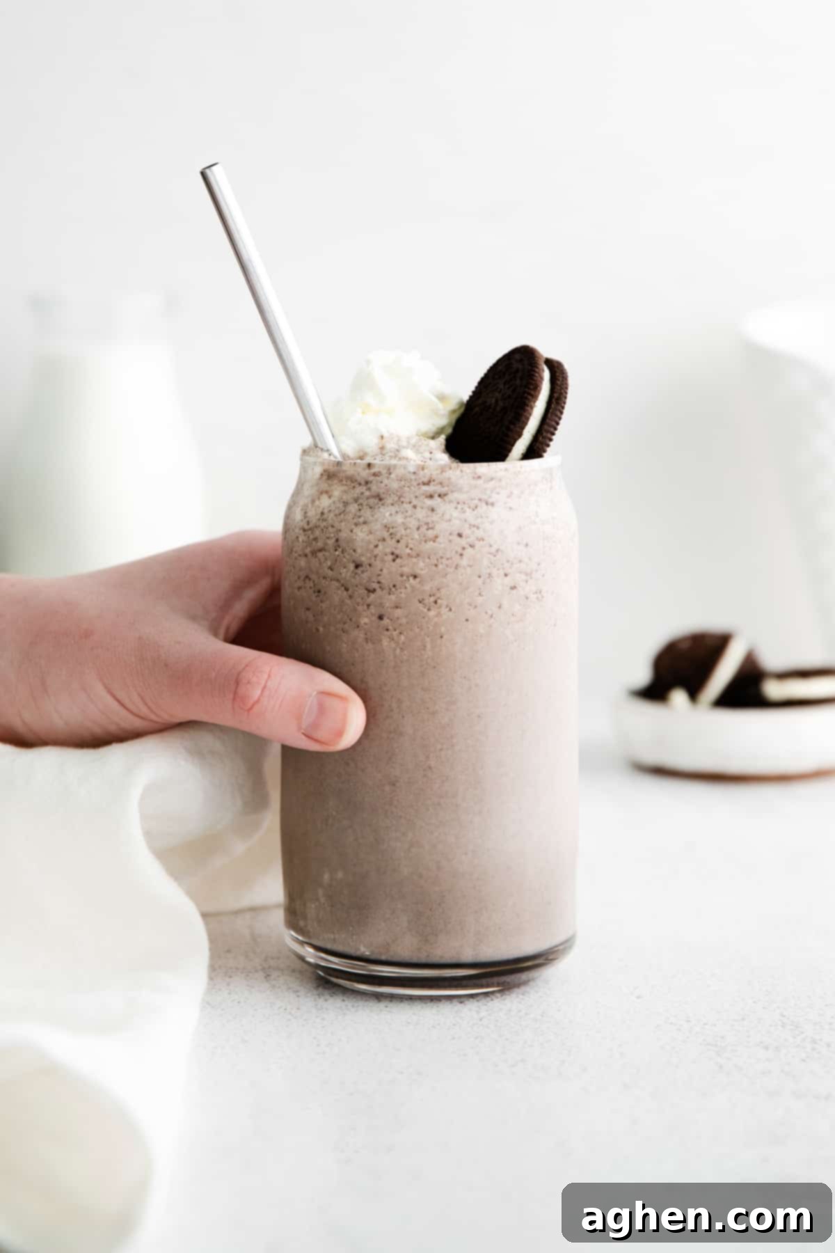 A hand reaching in to pick up a tall glass of Boozy Oreo Milkshake, perfectly blended and garnished with whipped cream and an OREO cookie.