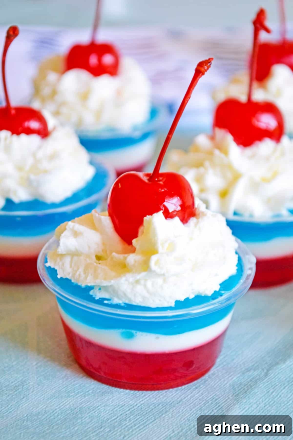 Layers of red, white, and blue gelatin topped with whipped cream and a maraschino cherry in a small clear plastic cup