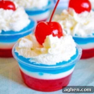 Layers of red, white, and blue gelatin topped with whipped cream and a maraschino cherry in a small clear plastic cup