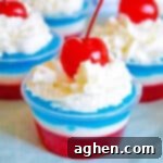 Layers of red, white, and blue gelatin topped with whipped cream and a maraschino cherry in a small clear plastic cup