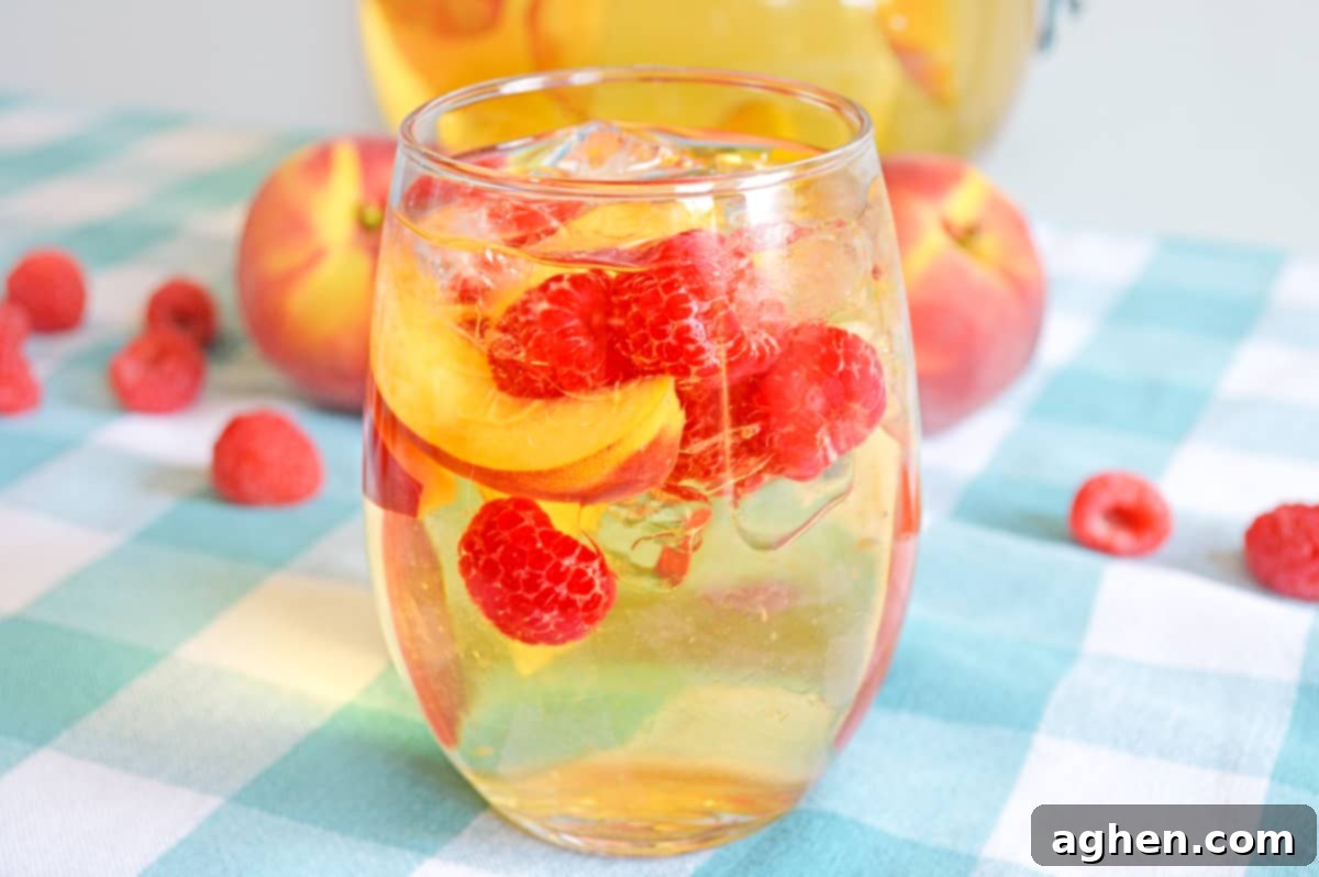 Refreshing Peach Sangria 2 White peach sangria with fresh peaches and raspberries in a stemless wine glass with whole peaches and raspberries in background