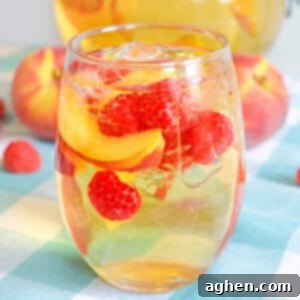 Refreshing Peach Sangria 5 White peach sangria with fresh peaches and raspberries in a stemless wine glass with whole peaches in background