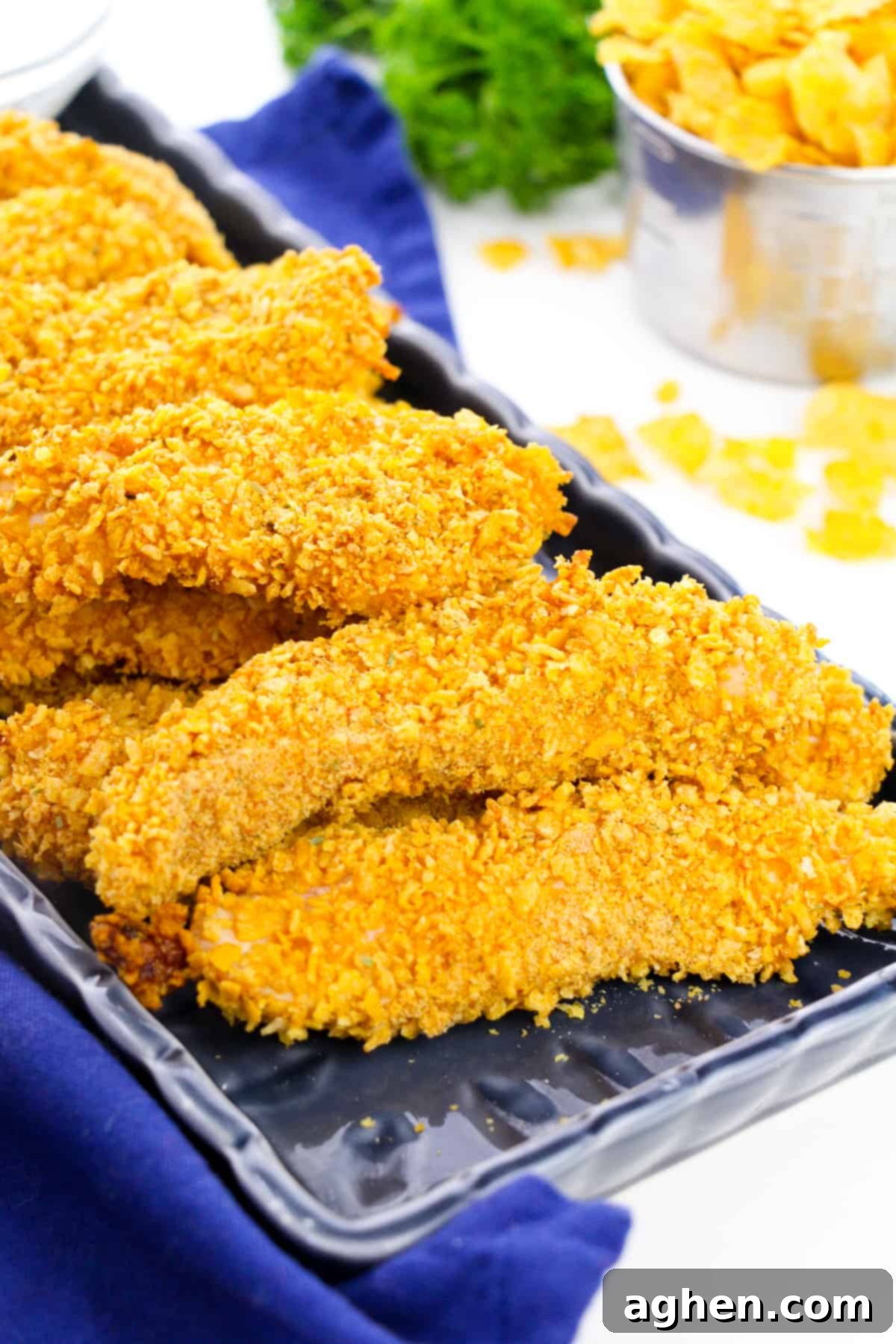 Golden-brown crispy cornflake chicken tenders on a black plate, garnished with fresh herbs, with crushed cornflakes in the background.