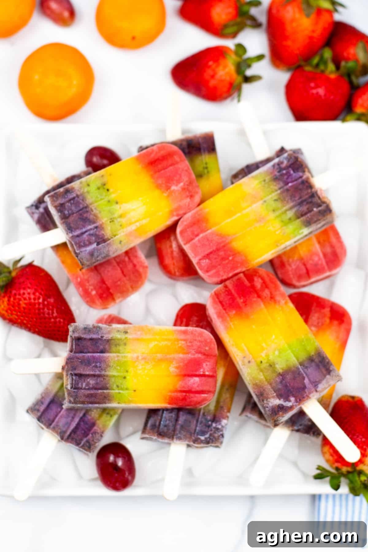 Bright and colorful rainbow fruit popsicles served on a bed of ice on a white plate. Mandarin oranges, strawberries, and grapes surround the plate.