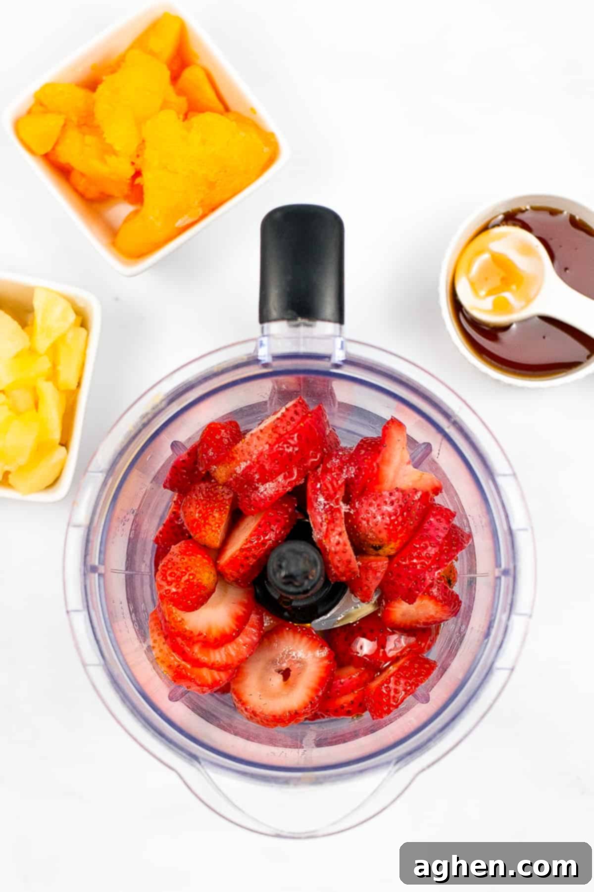 Sliced strawberries in blender with bowls of pineapple chunks, mandarin orange slices, and honey around it