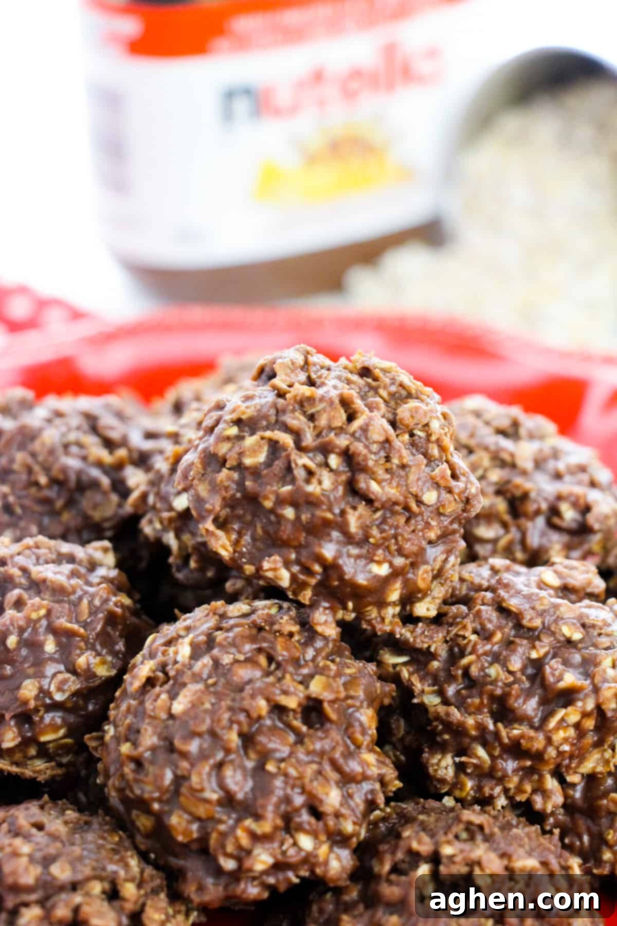 A close-up shot of thick, rich chocolate Nutella no-bake cookies, generously piled on a red plate, with a jar of Nutella and a measuring scoop of quick oats in the soft background.