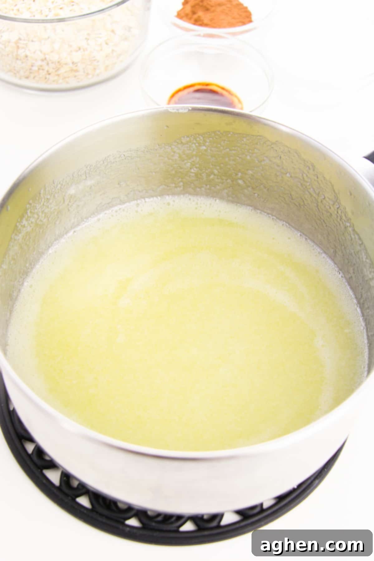 A saucepan on a stovetop, containing melted butter, milk, and white granulated sugar, actively boiling, with additional cookie ingredients visible in the soft focus background.