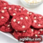 Red velvet cookies with white chocolate chips piled on white serving platter with bowl of white chocolate chips in the background