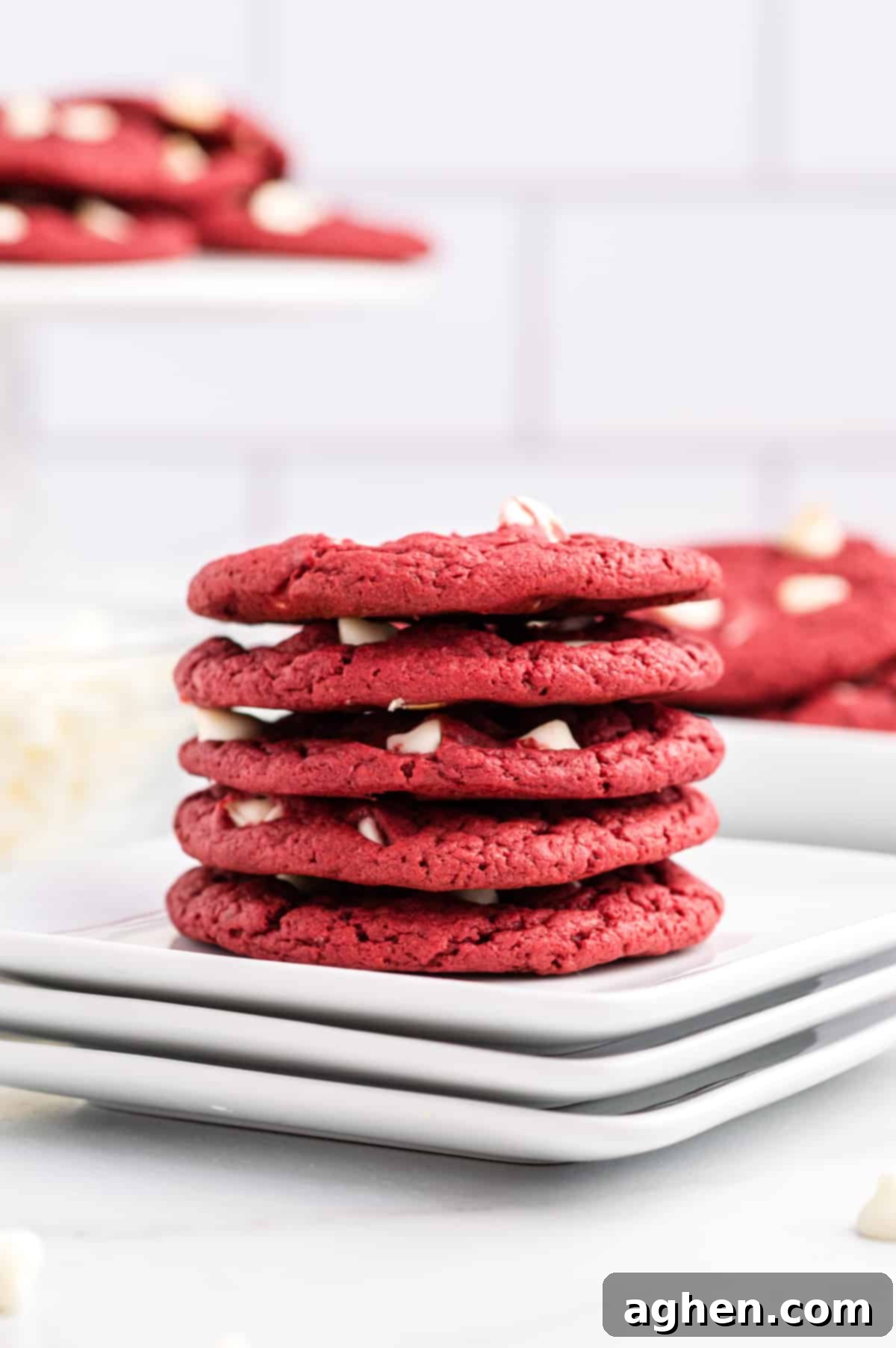 Red velvet white chocolate cookies stacked on white plates. More cookies can be seen in background.