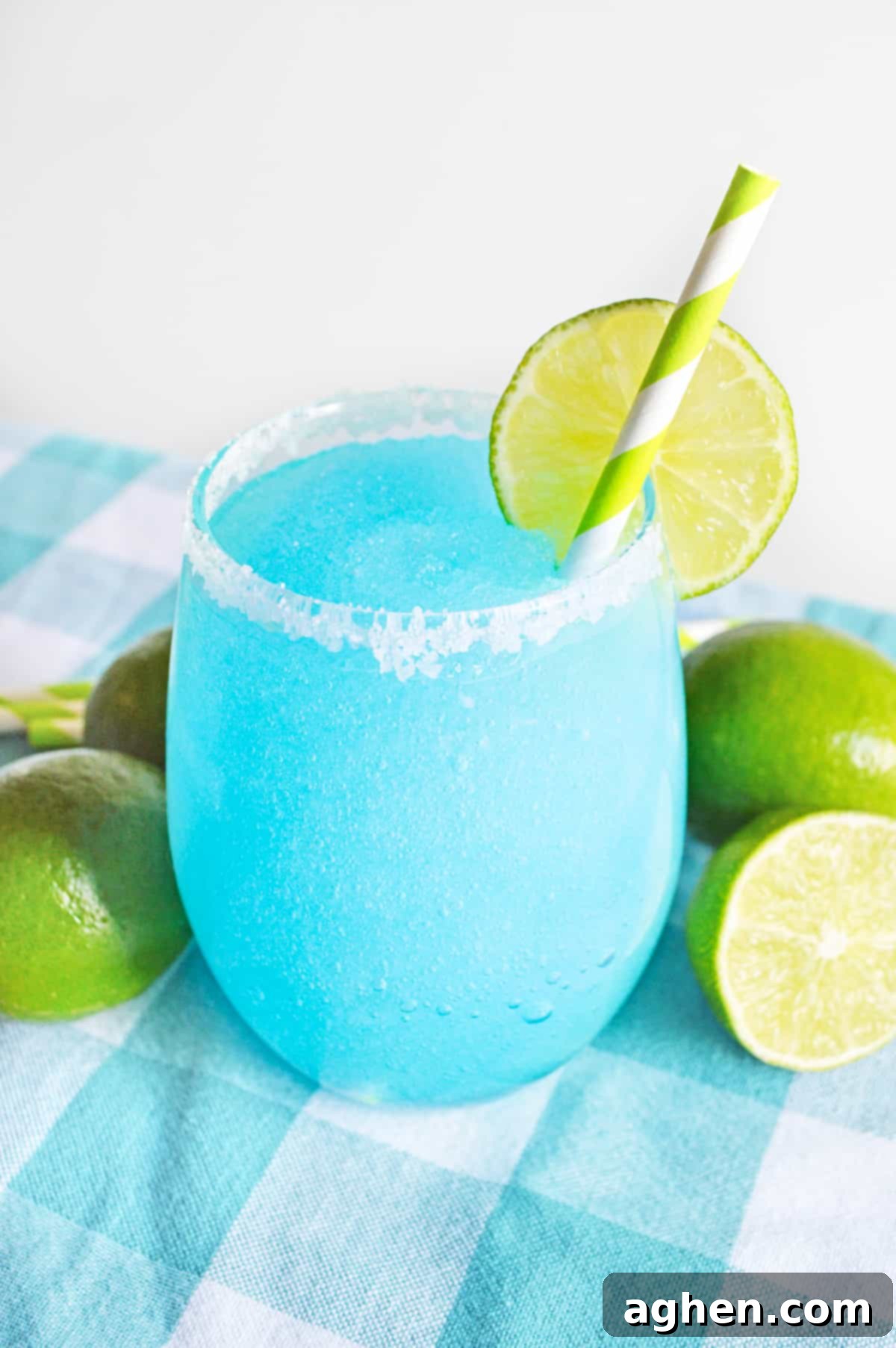 Azure Bliss Margaritas 2 Bright blue frozen margarita garnished with lime and served in a glass rimmed with coarse sugar. Limes are next to glass on blue tablecloth.
