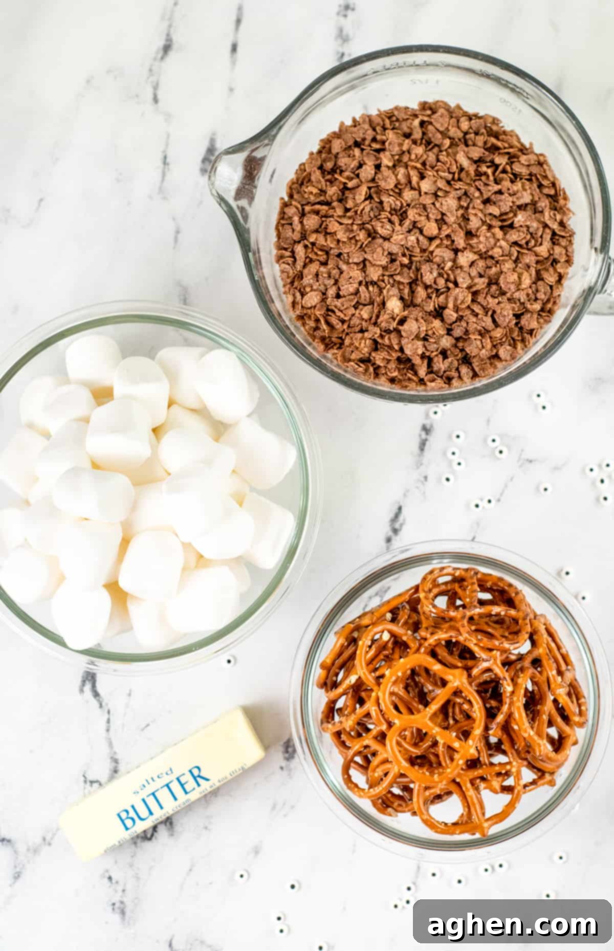 Frightfully Delicious Spider Bites 4 Ingredients for Spider Krispies Treats on a countertop: a bowl of marshmallows, a bowl of chocolate krispy cereal, a stick of butter, and a bowl of thin pretzels.