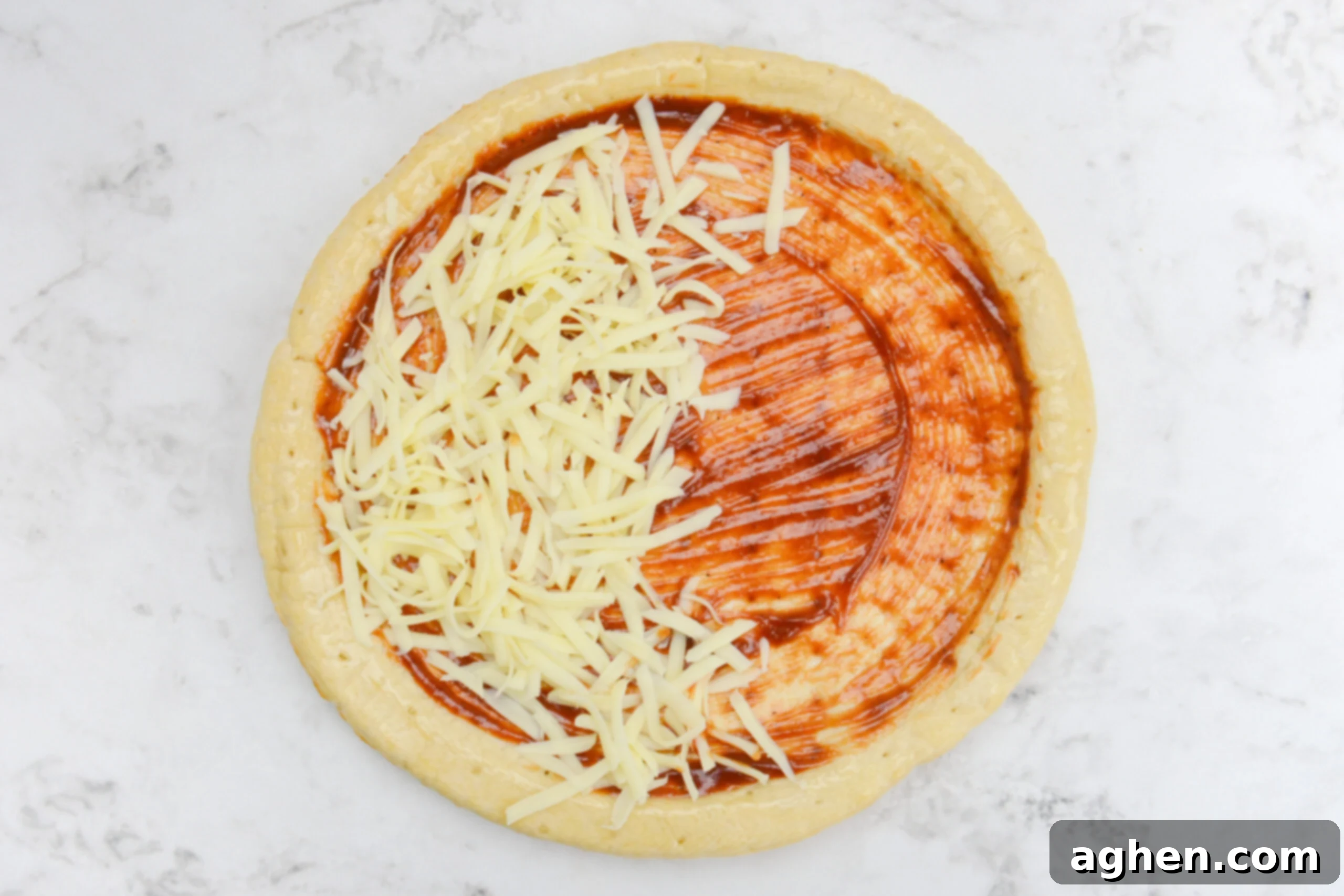An overhead view of the pizza crust with a layer of sauce and half of the crust covered in shredded mozzarella.