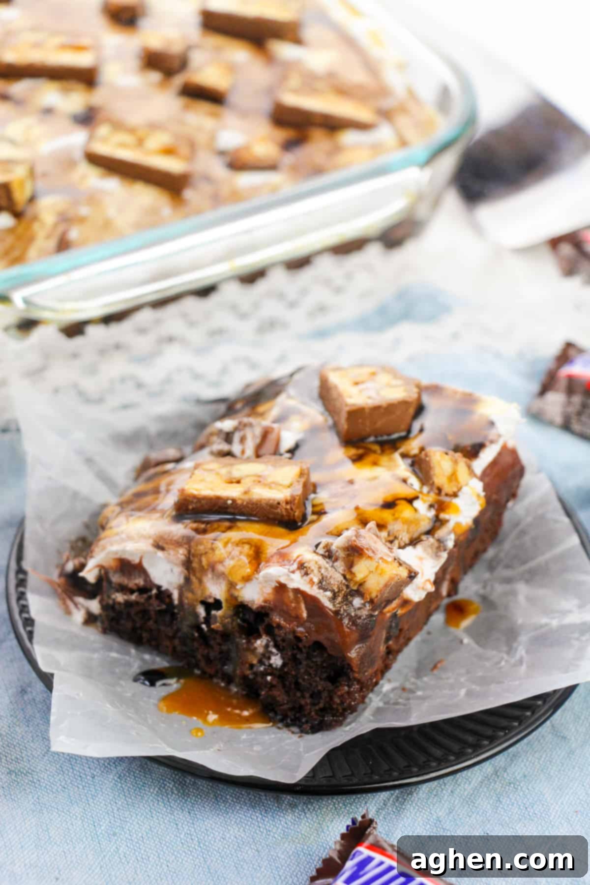 Close-up of a moist Snickers poke cake slice showing layers of chocolate cake, caramel, chocolate pudding, cool whip, peanuts, and chopped Snickers candy bars.
