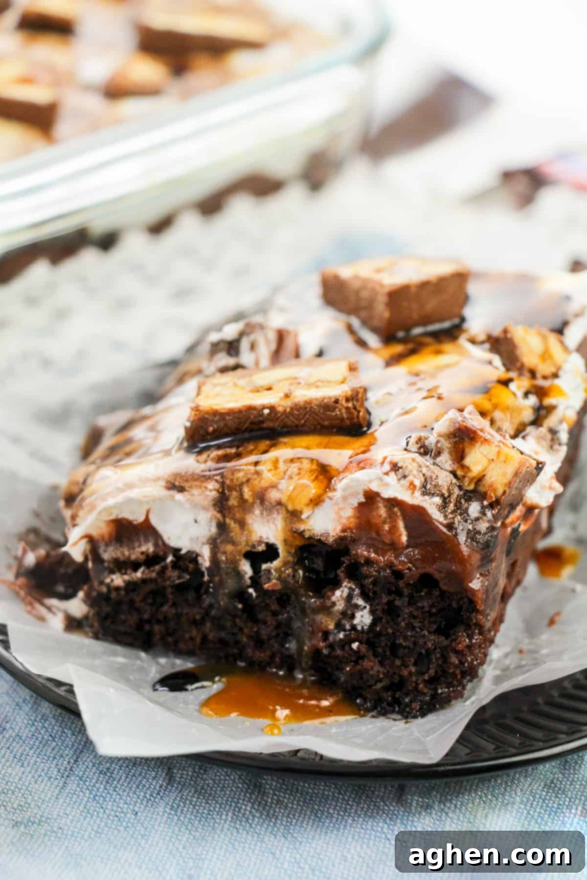 A slice of Snickers poke cake on a plate, with a background of the full cake, showcasing the layers and drizzles of chocolate and caramel sauces.