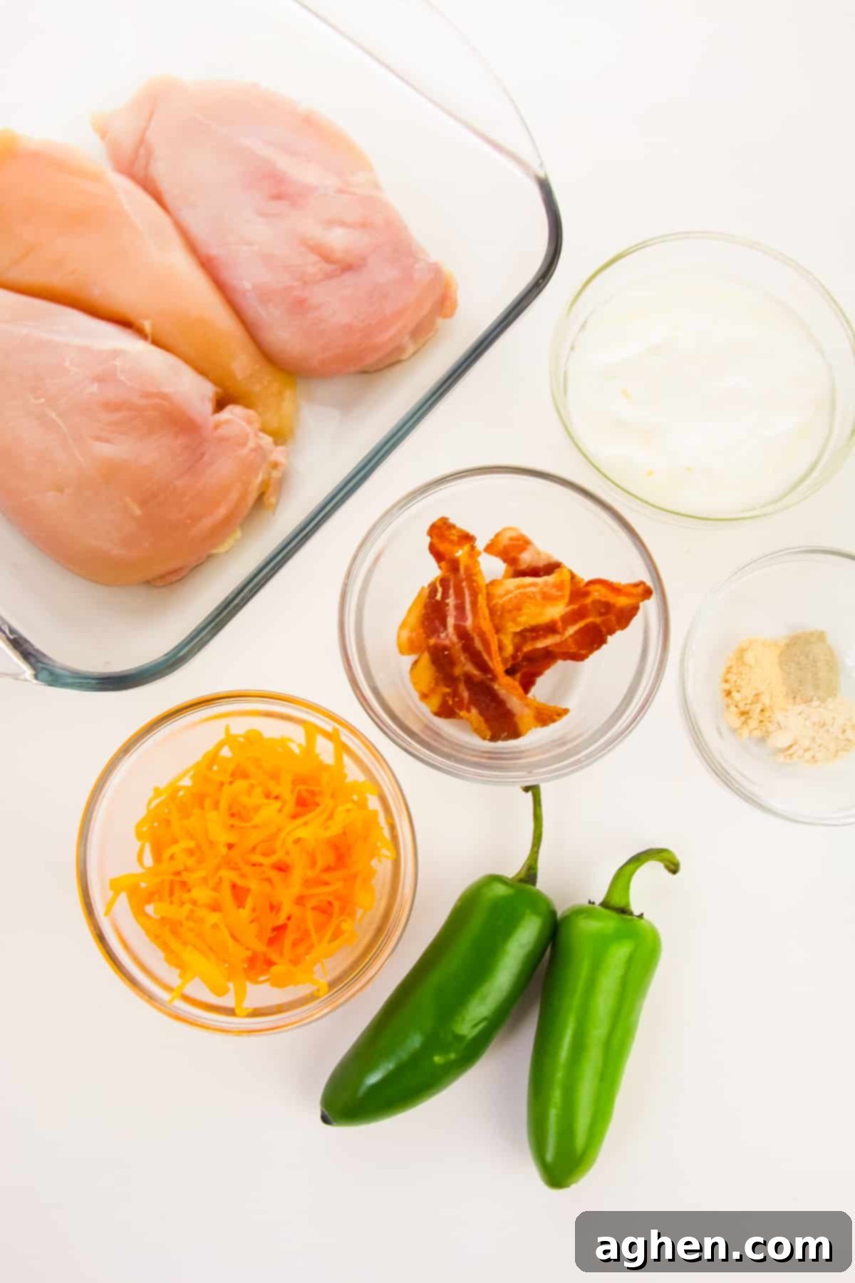Ingredients for Jalapeño Popper Chicken: three chicken breasts in a baking dish, two fresh jalapeño peppers, and glass bowls containing shredded cheese, bacon bits, mayonnaise/sour cream mixture, and various seasonings.