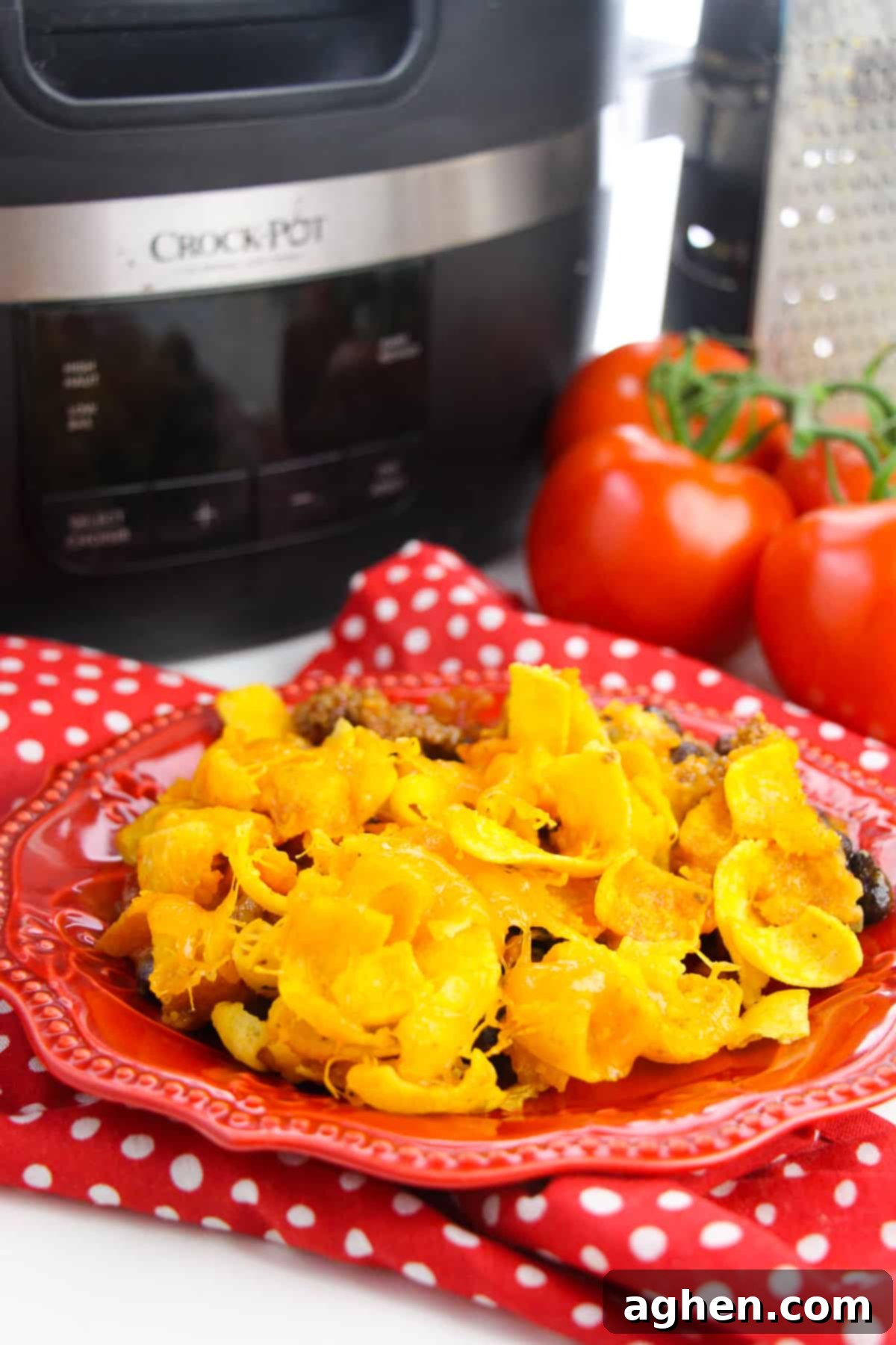 Close-up of Crockpot Frito Pie served on a red plate, with a slow cooker, fresh tomatoes, and cheese grater blurred in the background, highlighting the rich texture and vibrant colors of this comforting dish.