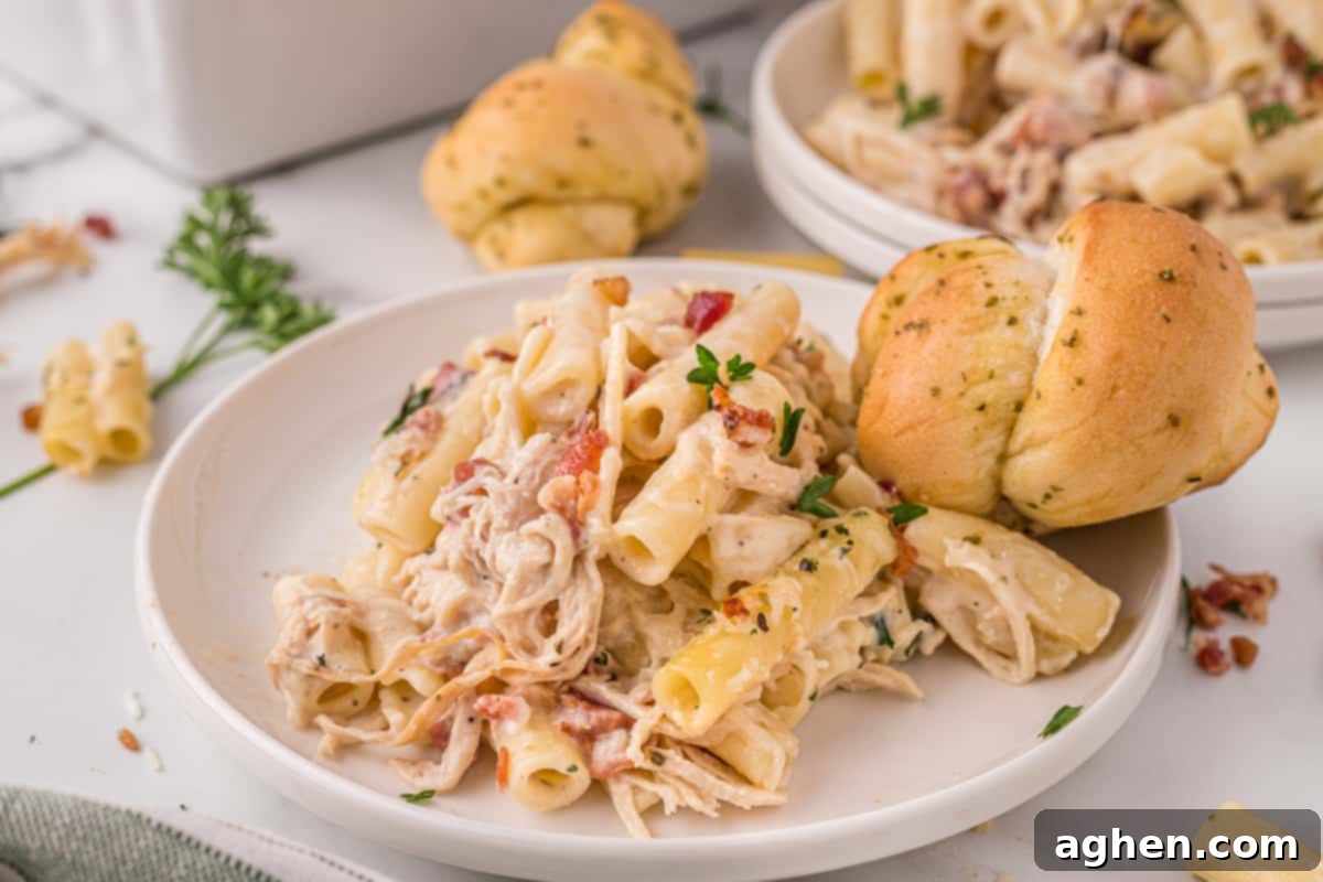 Serving of cheesy chicken alfredo pasta bake on a white plate with a roll and an additional plate and roll in the background