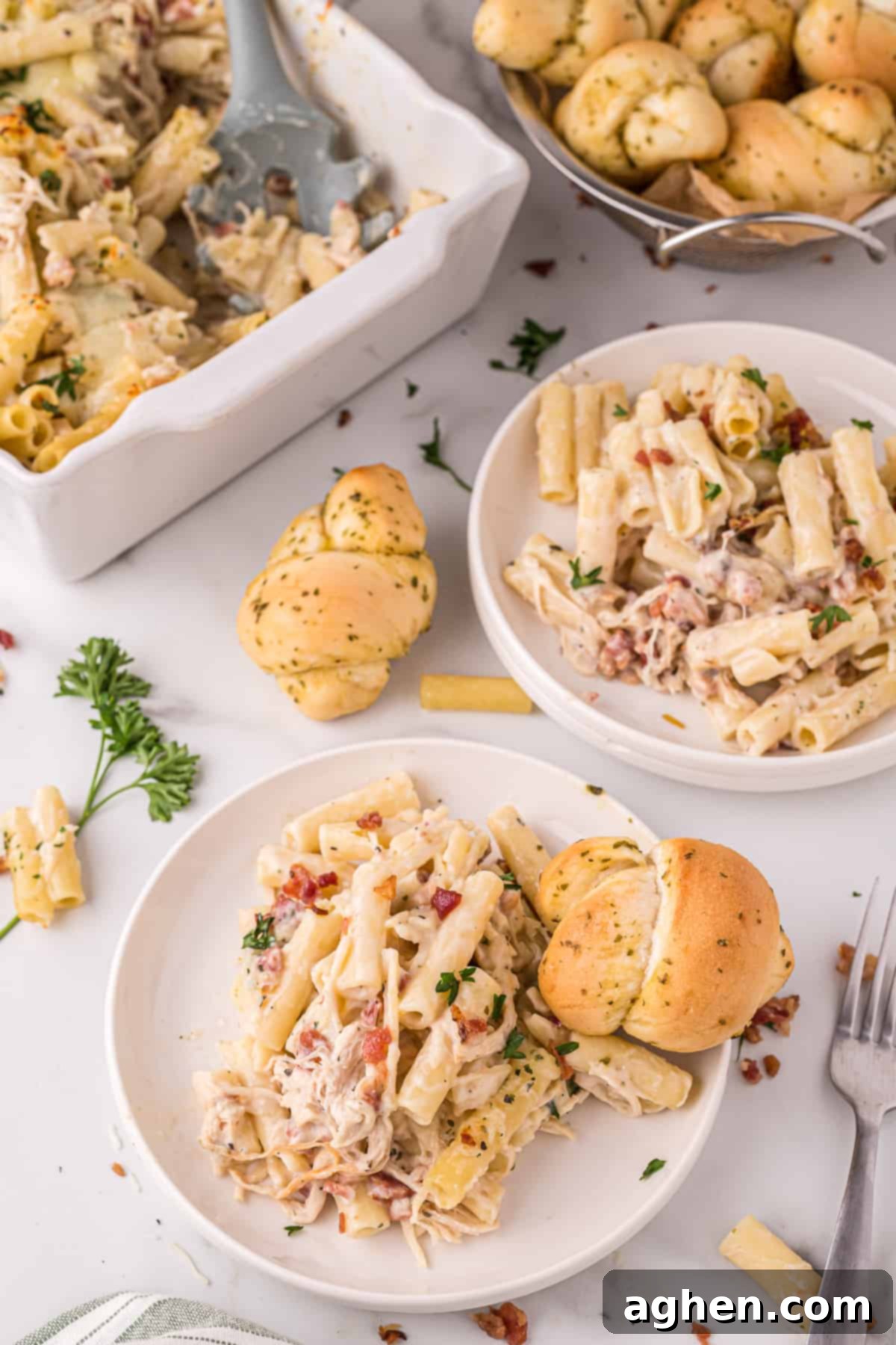 Serving of chicken bacon ziti pasta bake on a white plate with a garlic knot and an additional plated portion, more rolls, and casserole dish in the background 