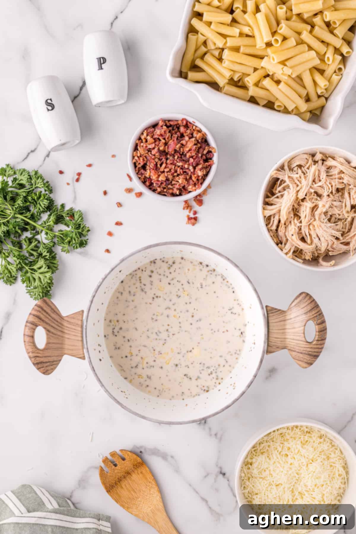 Alfredo sauce in large pot with ziti, chicken, cheese, parsley, bacon and other ingredients around it