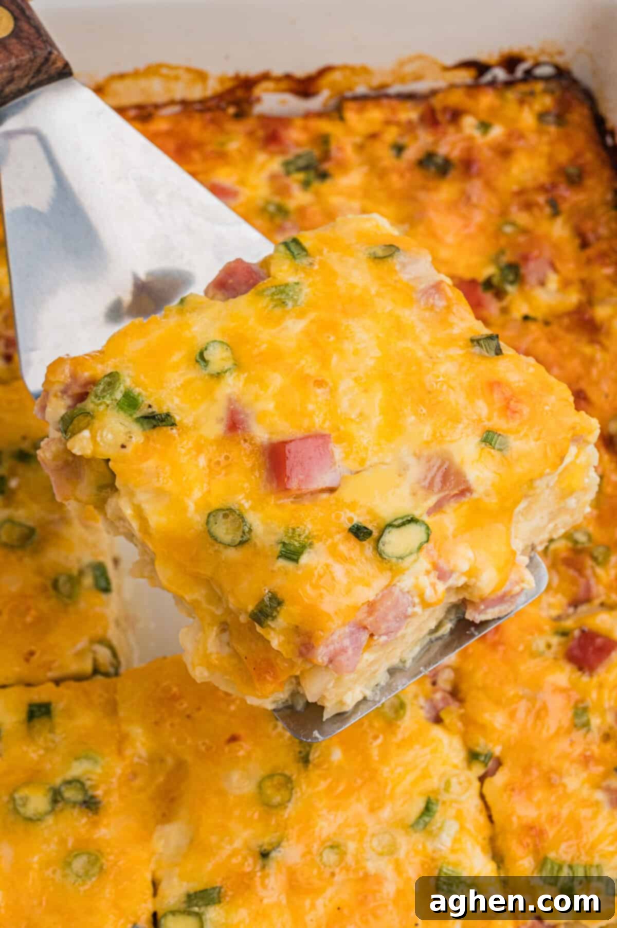 Close-up of a farmers breakfast casserole piece held on a metal spatula, highlighting chunks of ham, bits of green onion, and golden melted cheese.