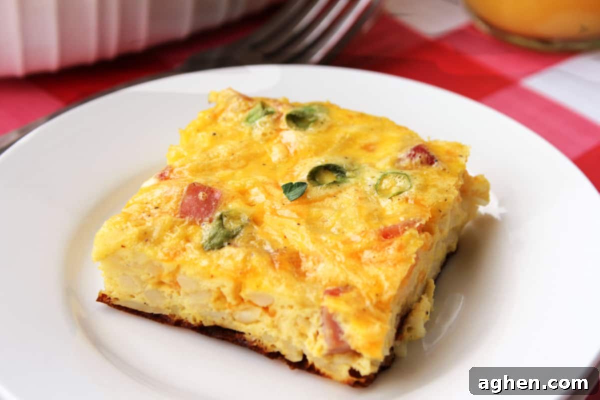 Square slice of breakfast casserole with hash browns, ham, eggs, and green onion served on a white plate with a fork next to it