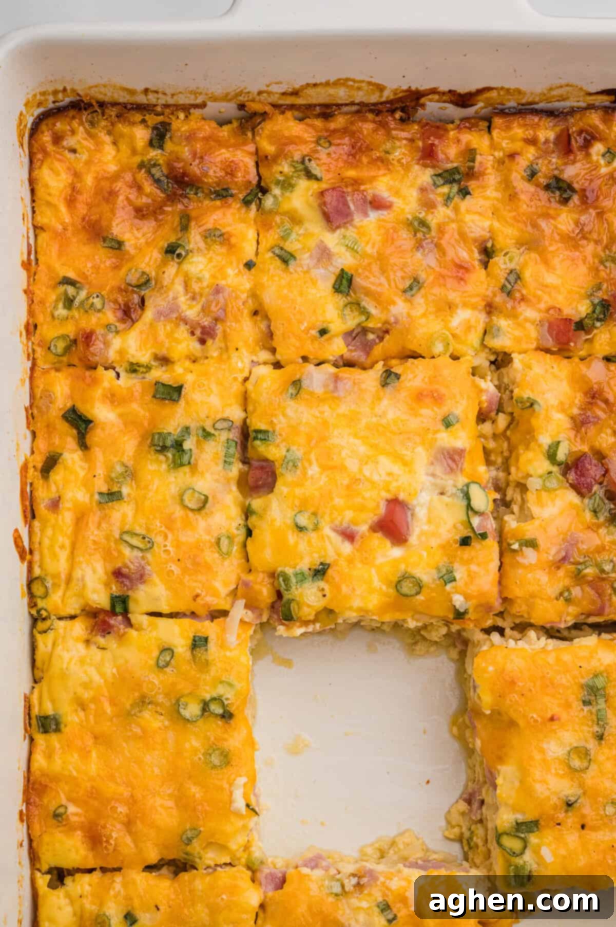 Overhead view of a white baking dish with a sliced breakfast casserole, featuring melted cheese, ham, and green onions, with the center piece removed.