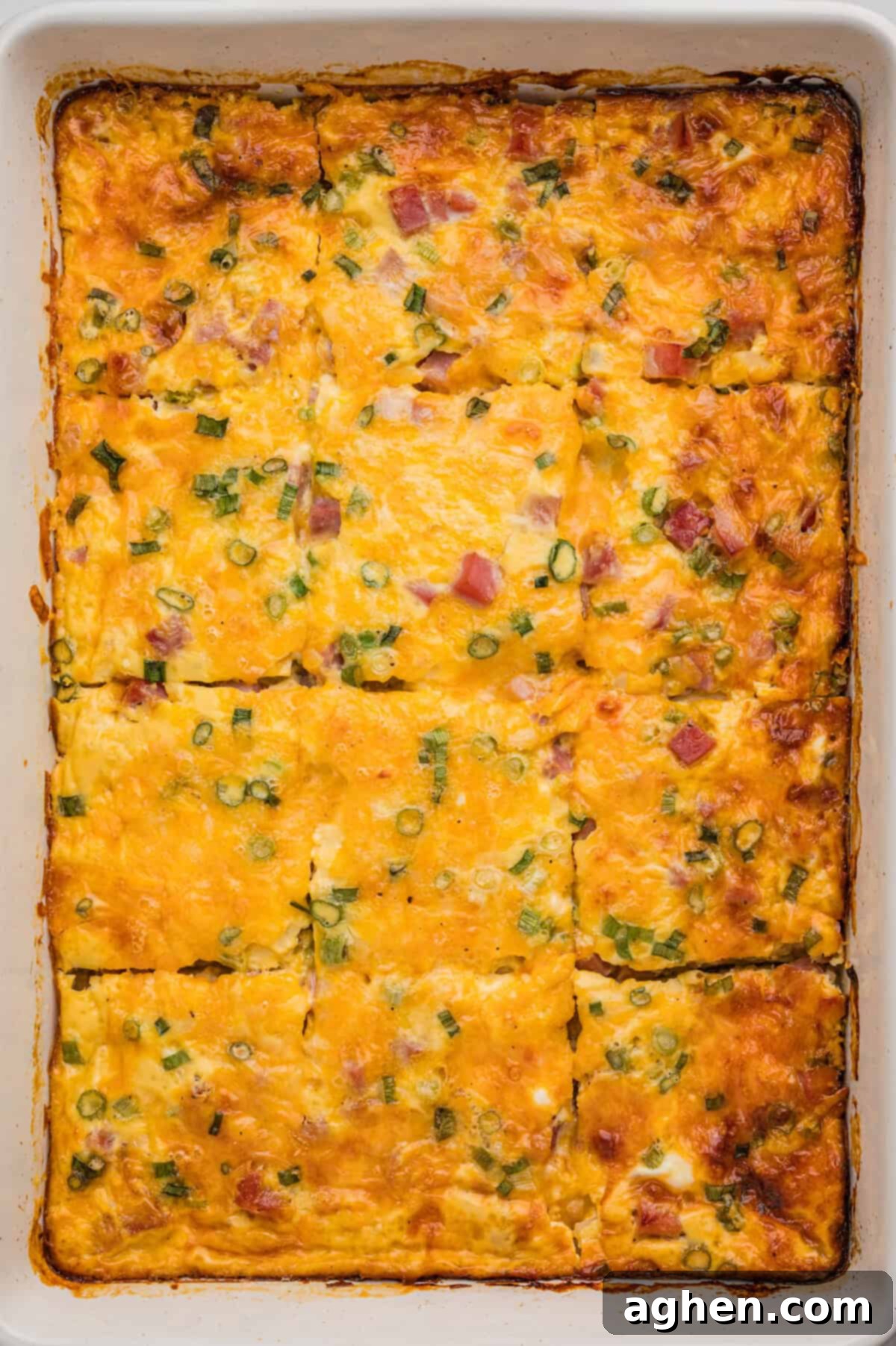 A fully baked farmer's casserole in a white dish, cut into rectangular slices with golden melted cheese, diced ham, and green onion visible on top.