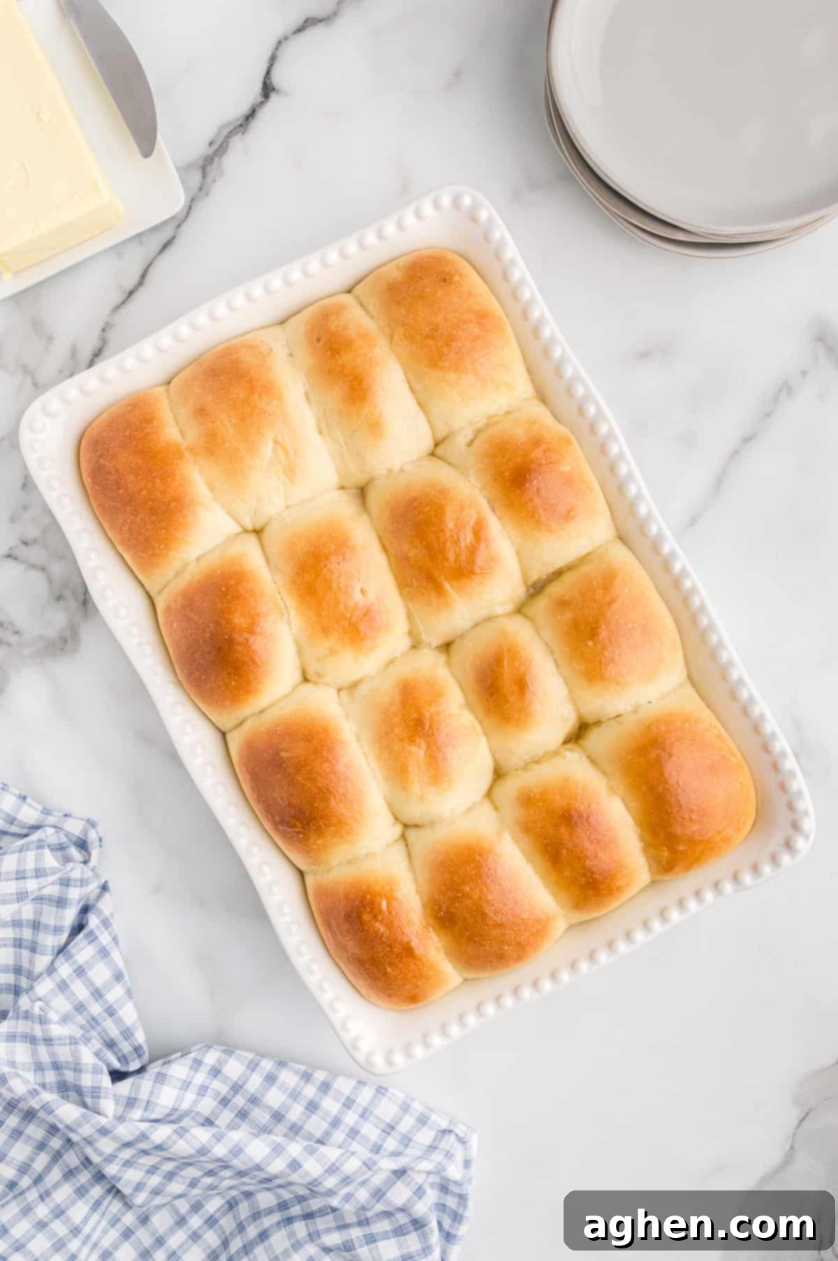 16 golden brown potato dinner rolls in white baking dish with plates, linen, and butter around them