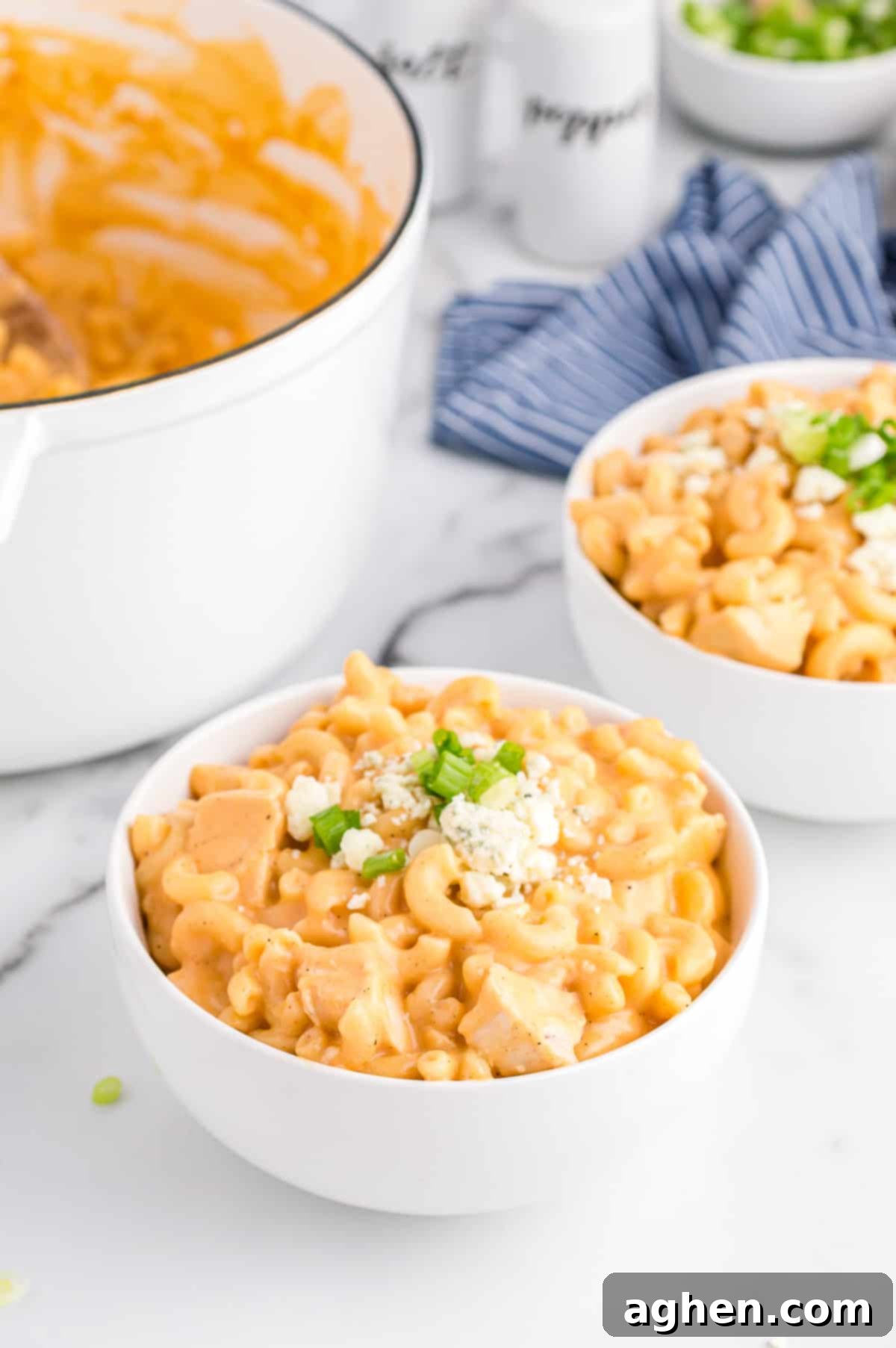 Buffalo Chicken Mac and Cheese in white bowl topped with blue cheese and scallions. Another serving and a large pot with the rest of the pasta can be seen in background.