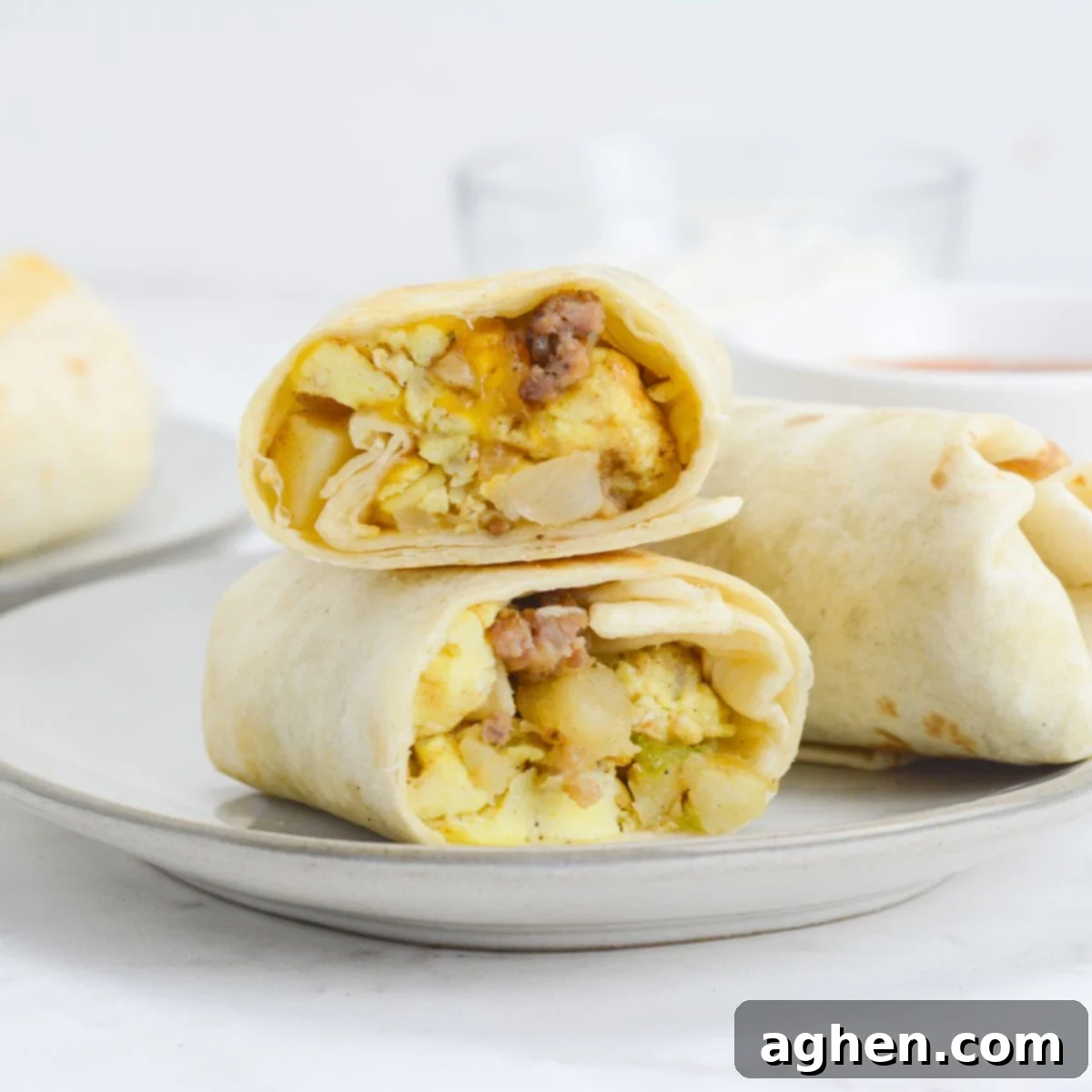 A breakfast burrito cut in half sits on a plate next to an uncut burrito. In the background there is a small bowl of hot sauce. 