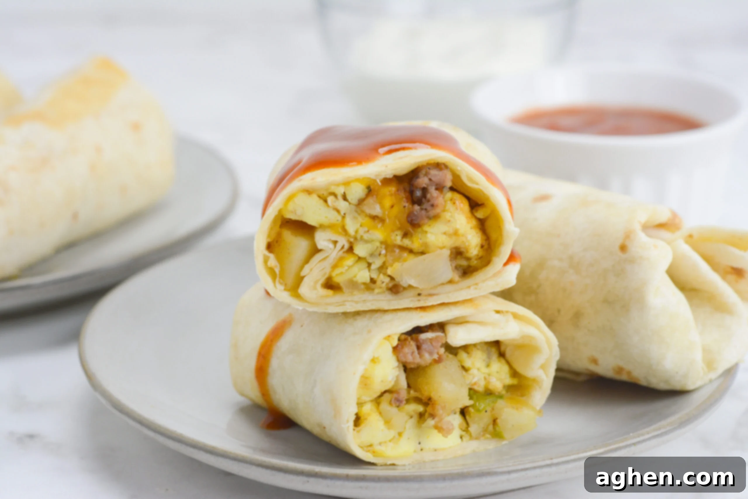 A burrito cut in half sits on half with hot sauce drizzled on top. An uncut burrito sits to the side and behind there are bowls with salsa and sour cream