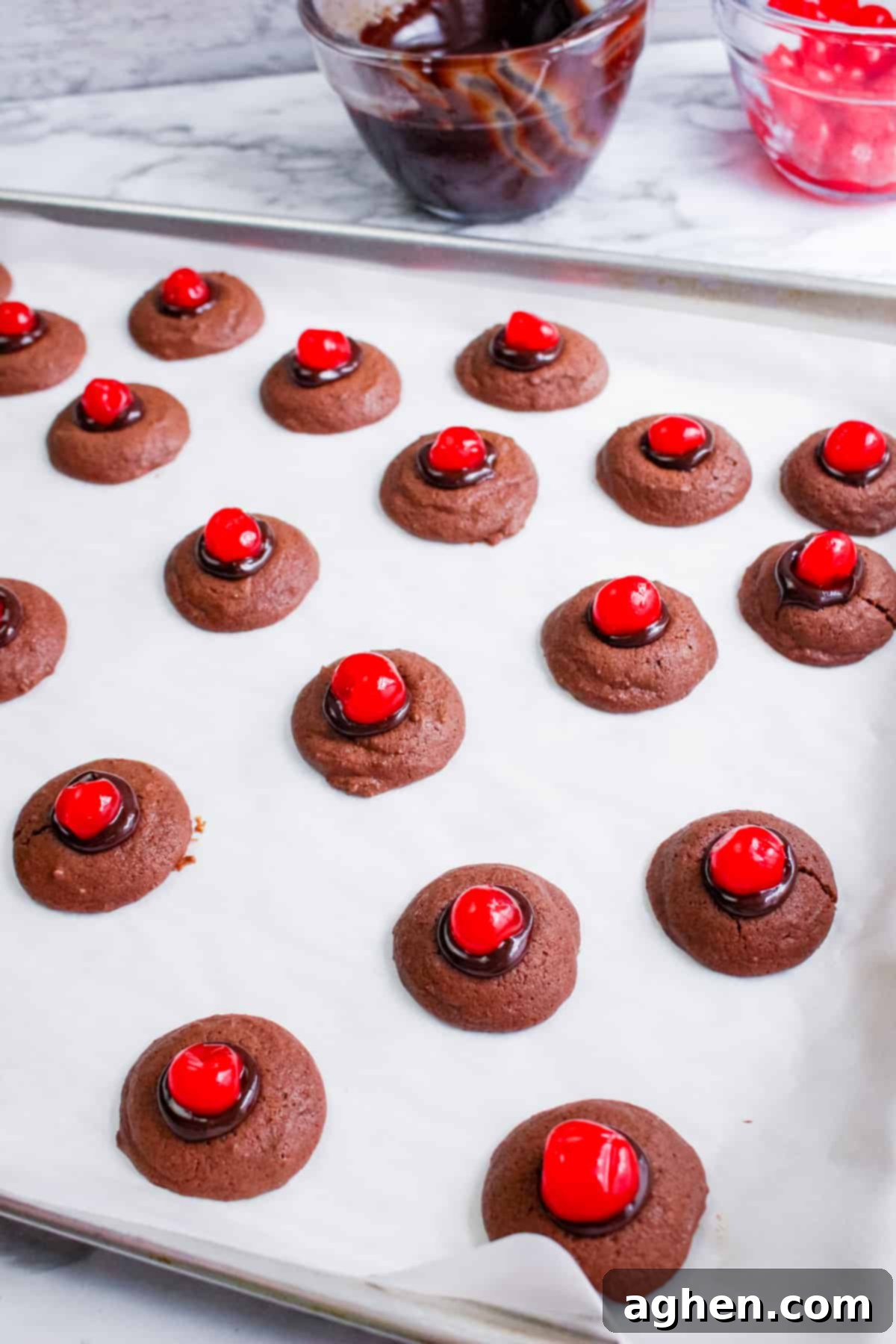 lined baking sheet with small chocolate thumbprint cookies topped with melted chocolate and maraschino cherries