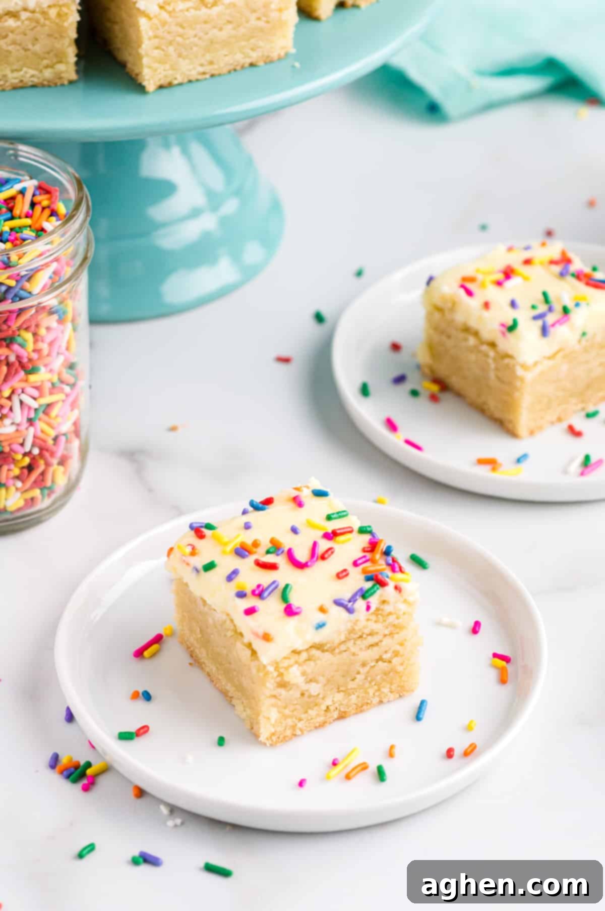Tender Frosted Sugar Cookie Bars 11 Two frosted sugar cookie bars with colorful rainbow sprinkles displayed on individual white plates. A jar of sprinkles and more baked bars are visible in the blurred background, creating a festive scene.