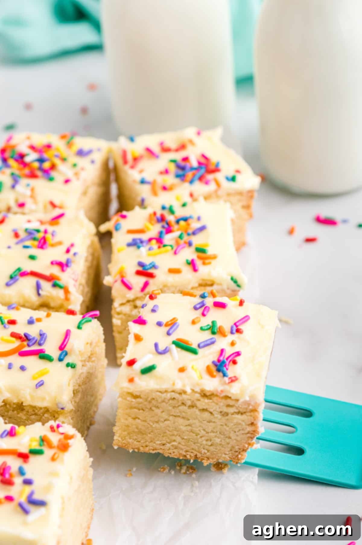 Tender Frosted Sugar Cookie Bars 4 A serving of sugar cookie bars with vanilla frosting and rainbow sprinkles, already cut into squares, being scooped onto a plate with a teal spatula. Glasses of milk are blurred in the background, suggesting a delightful pairing.