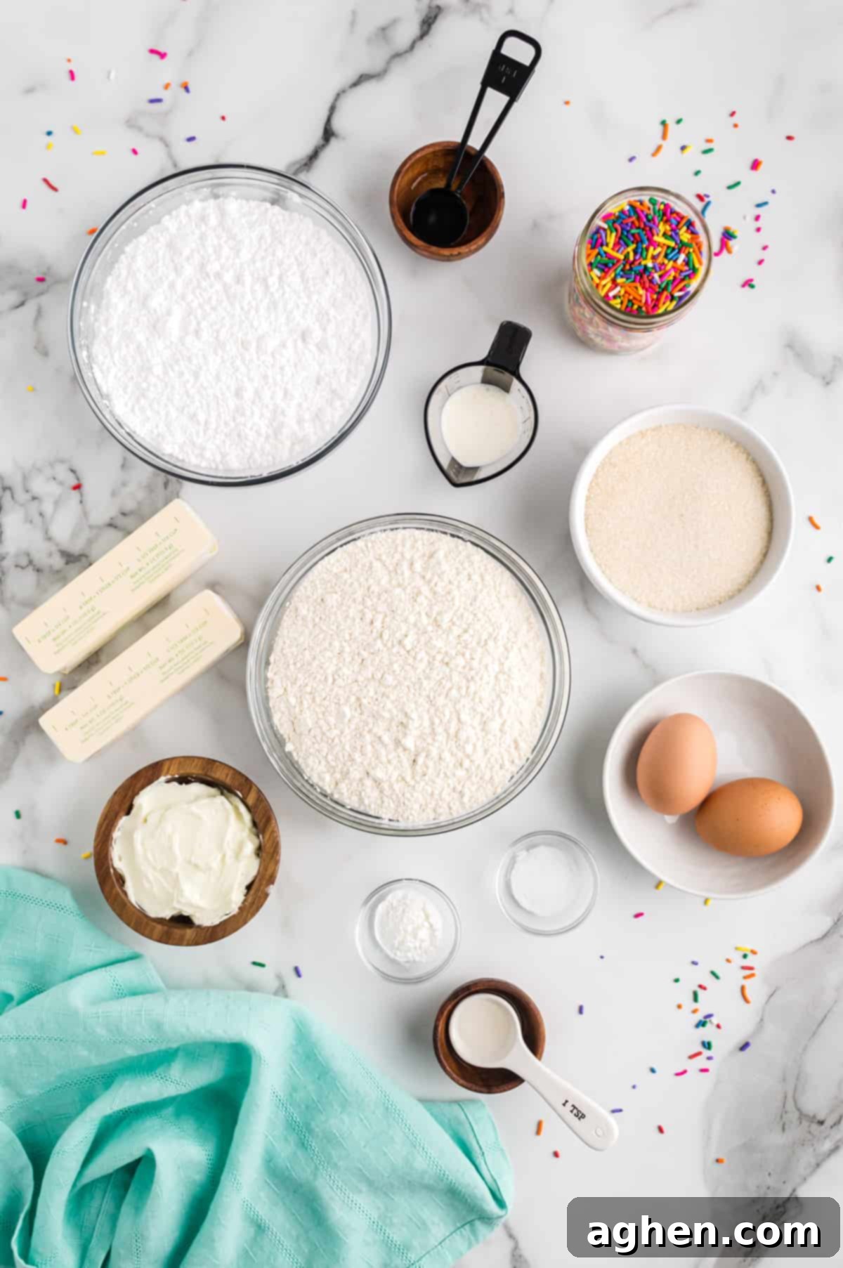 Tender Frosted Sugar Cookie Bars 5 A spread of ingredients on a kitchen countertop, including all-purpose flour, white granulated sugar, two sticks of softened butter, shortening, a bottle of clear vanilla extract, almond extract, two fresh eggs, salt, baking powder, powdered sugar, heavy cream, and a bowl of rainbow sprinkles.