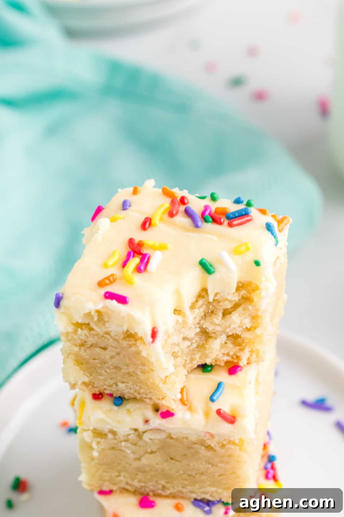 Tender Frosted Sugar Cookie Bars 10 Two frosted sugar cookie bars with sprinkles stacked on top of each other on a white surface. The top bar has a bite taken out, revealing its thick, tender, and chewy interior texture.