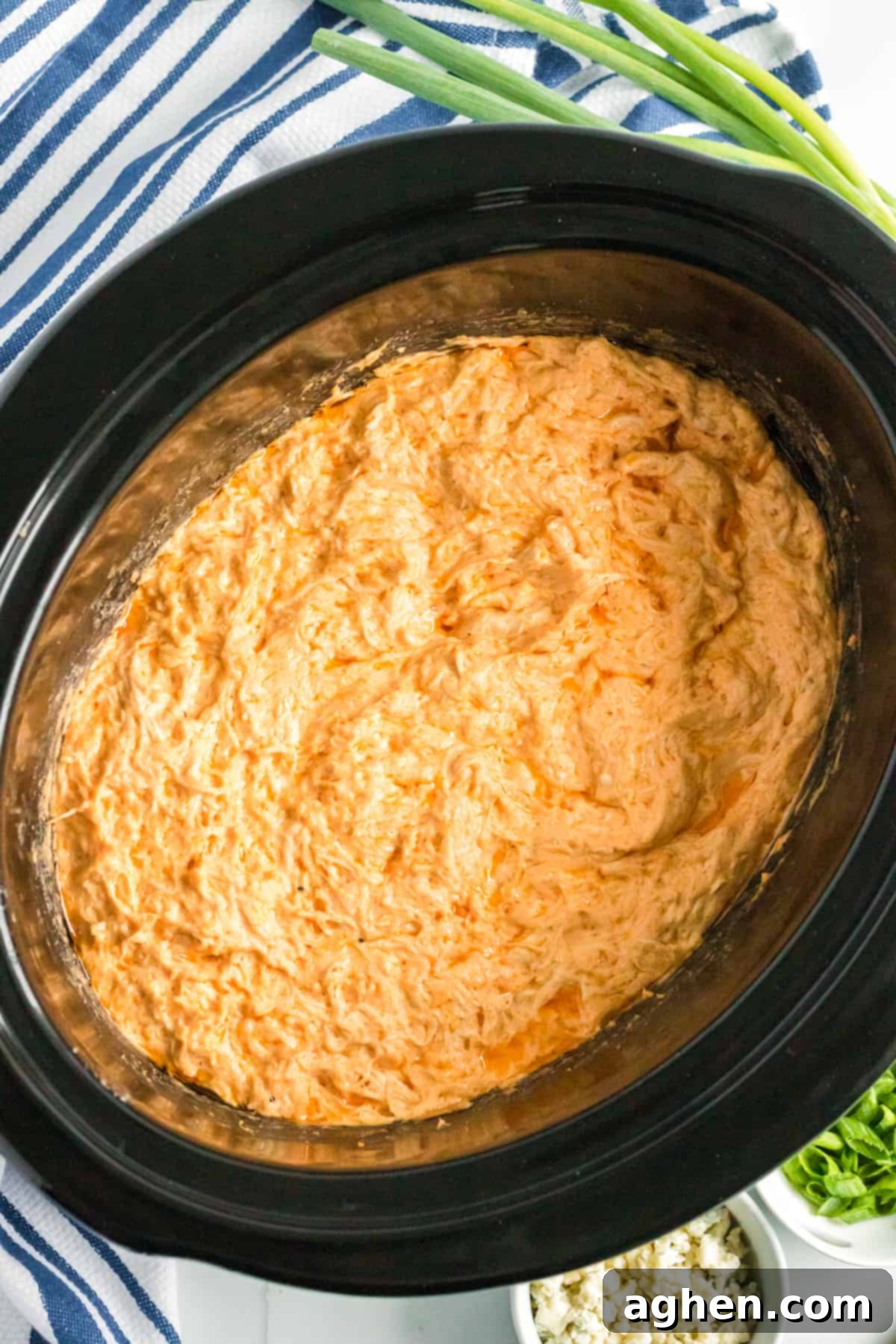 Buffalo chicken dip in the crockpot with green onions and crumbled blue cheese in the background.