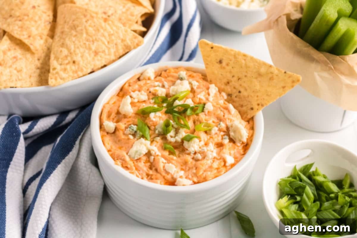 Slow Cooker Buffalo Chicken Dip in white bowl garnished with blue cheese and green onions. A tortilla chip is sticking out of the top.