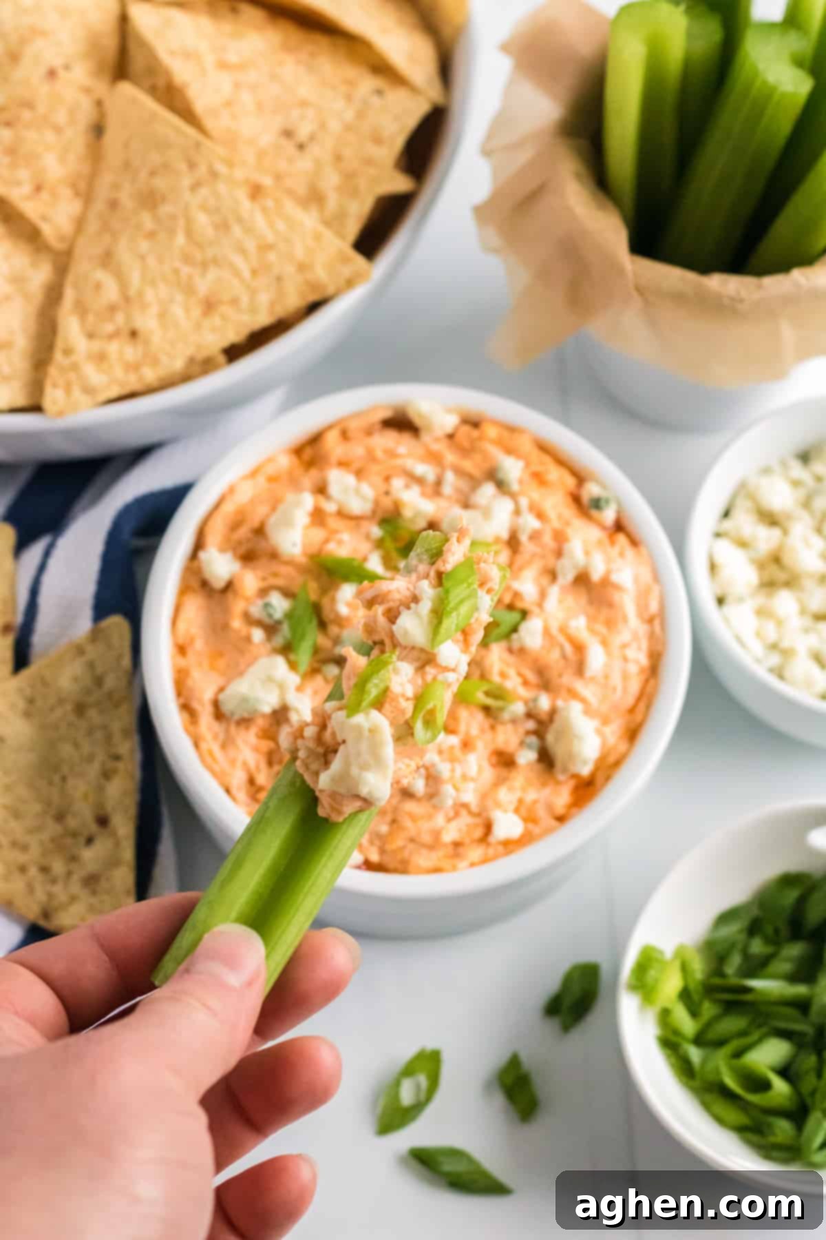 Celery stick being dipped in a bowl of cheesy buffalo chicken dip. Tortilla chips, more celery sticks, blue cheese crumbles, and scallions are in background.