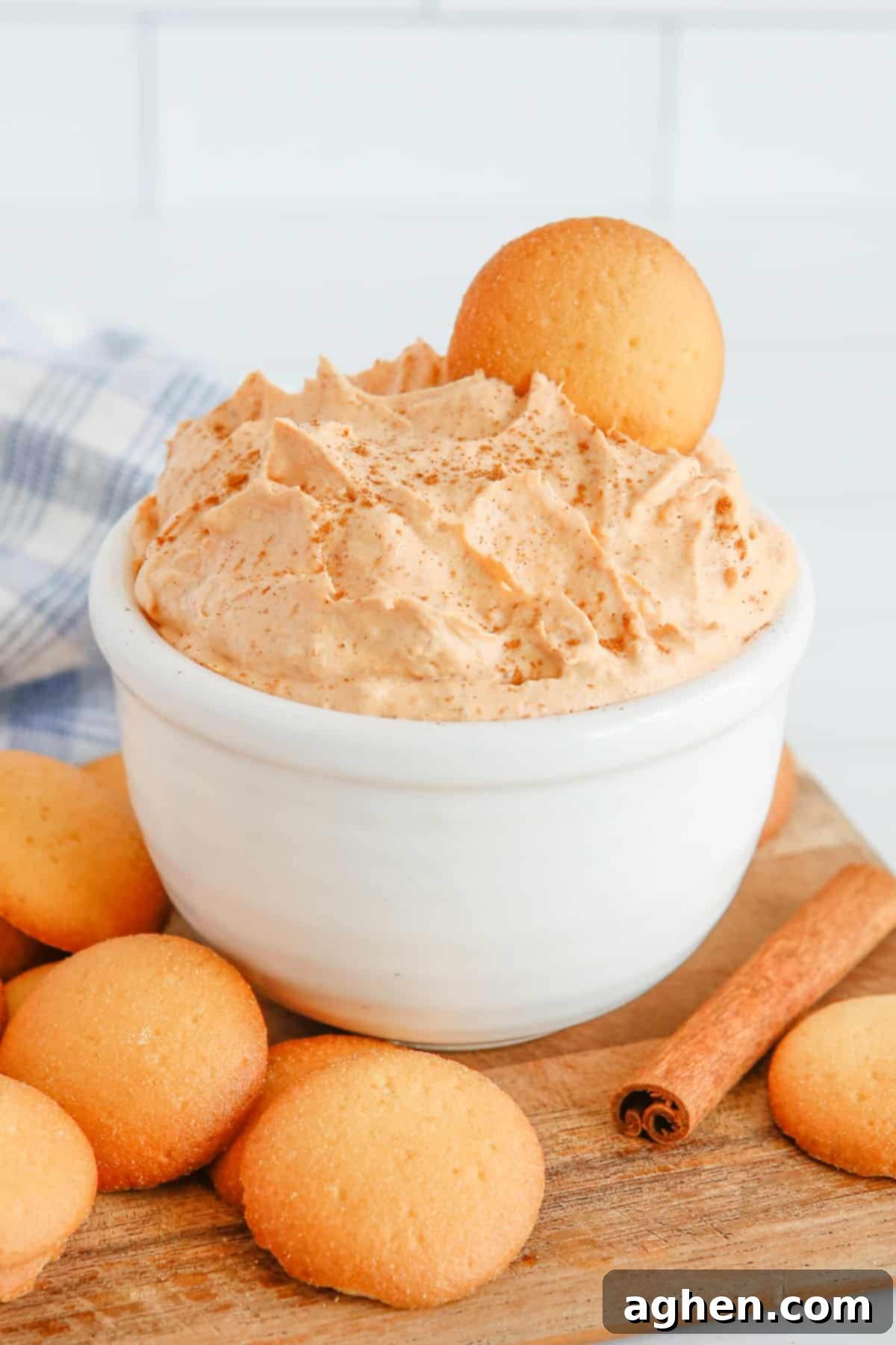 Pumpkin dip in white bowl with vanilla wafer sticking out of the top. Vanilla cookies and cinnamon sticks are on tabletop around the bowl