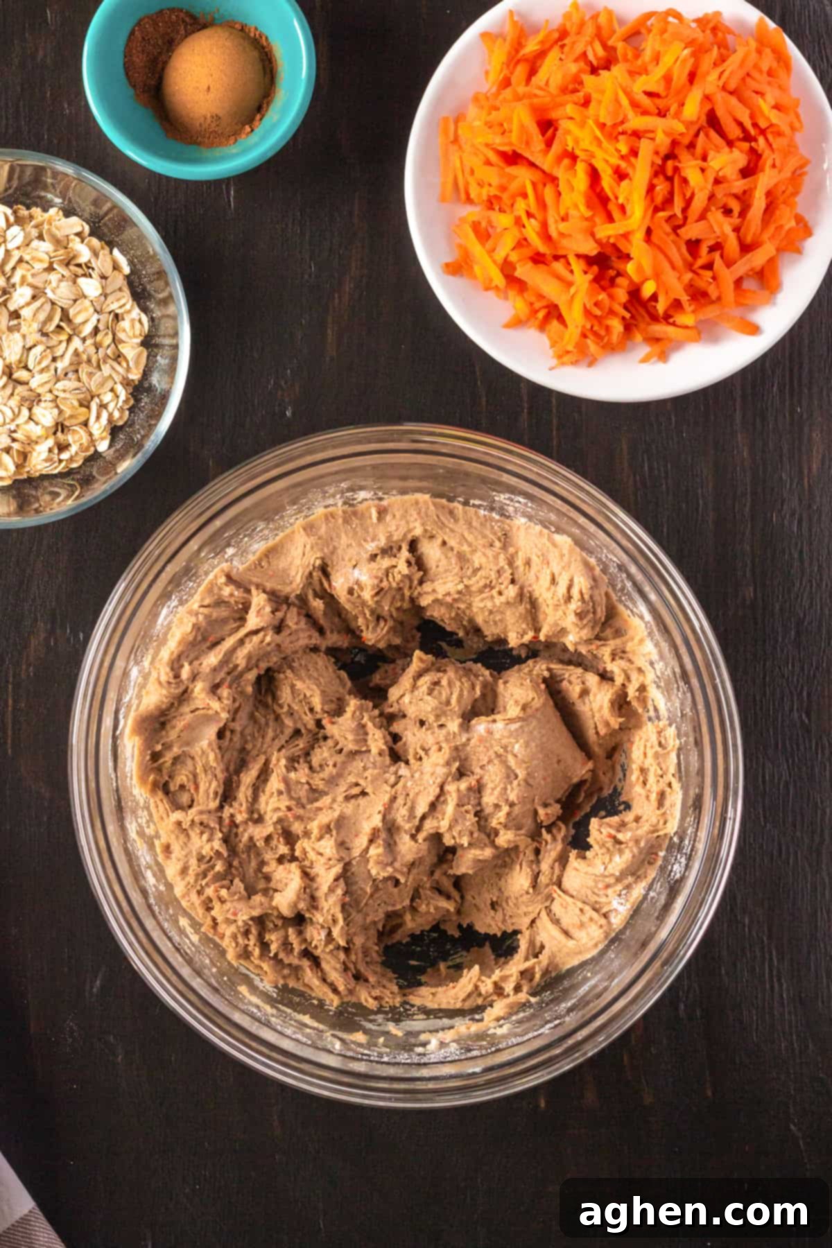 Cookie dough in a mixing bowl with shredded carrots, old-fashioned oats, and a small bowl of cinnamon and nutmeg sitting nearby, ready to be added.