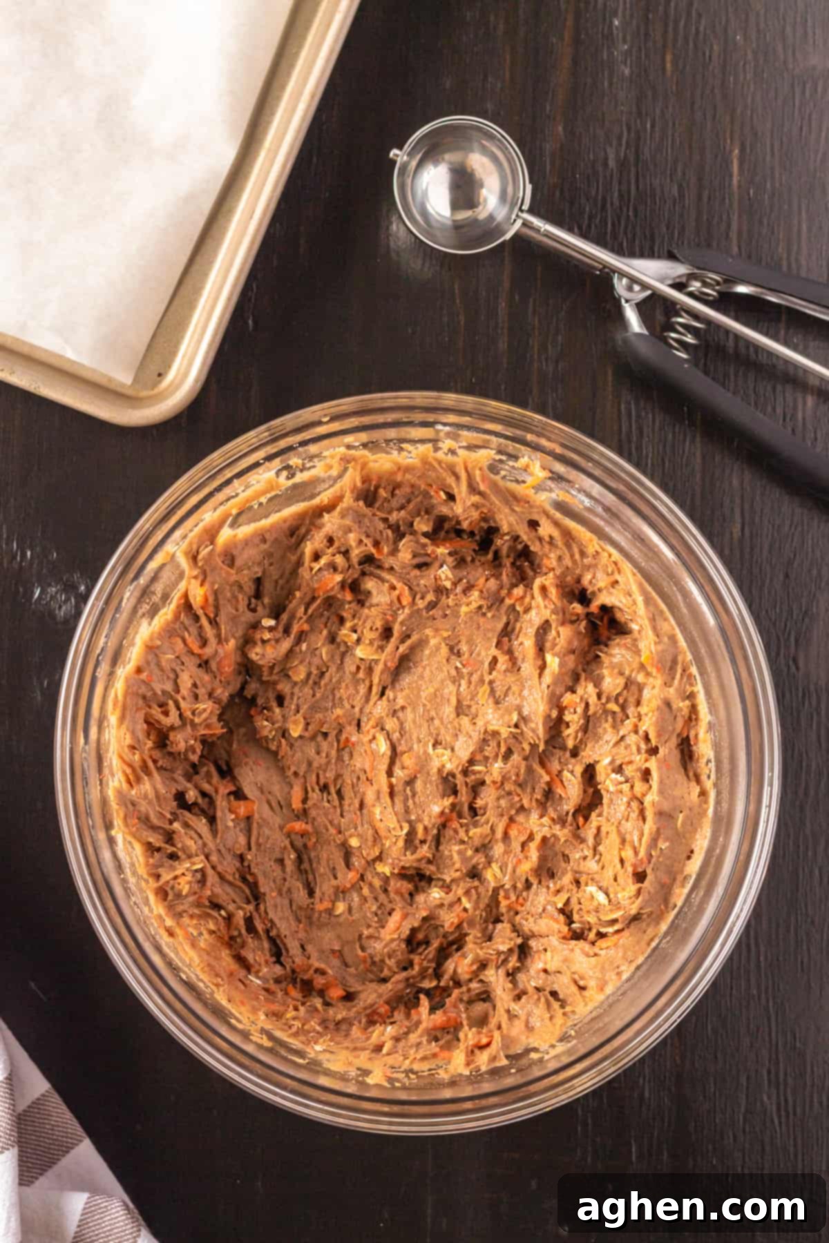 A bowl of carrot cake mix cookie dough, thick and studded with carrots and oats, with a cookie scoop and a parchment-lined baking sheet in the background.