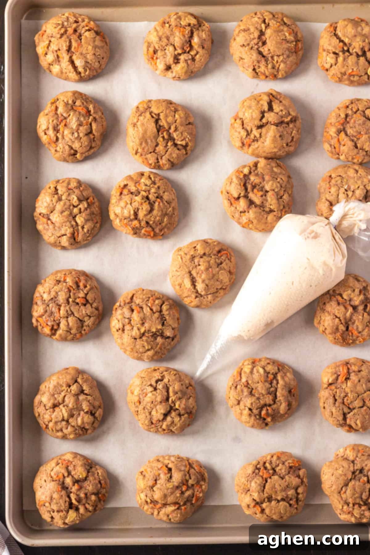 Warm, chewy carrot cake cookies cooling on a parchment-lined baking sheet, with a piping bag filled with cream cheese frosting positioned nearby.